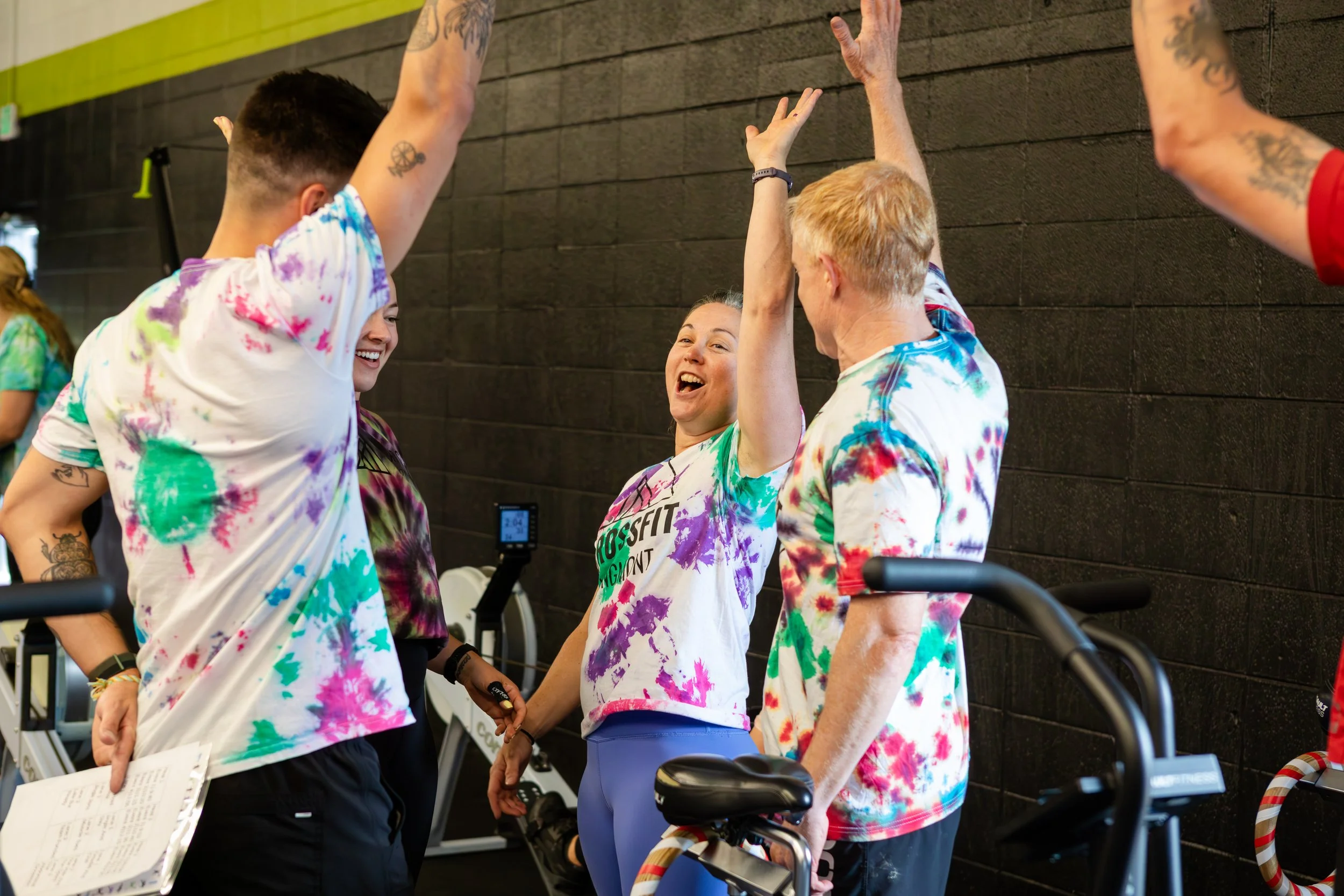 Group of four people in colorful tie-dye shirts standing together indoors, celebrating with high-fives, near exercise equipment.