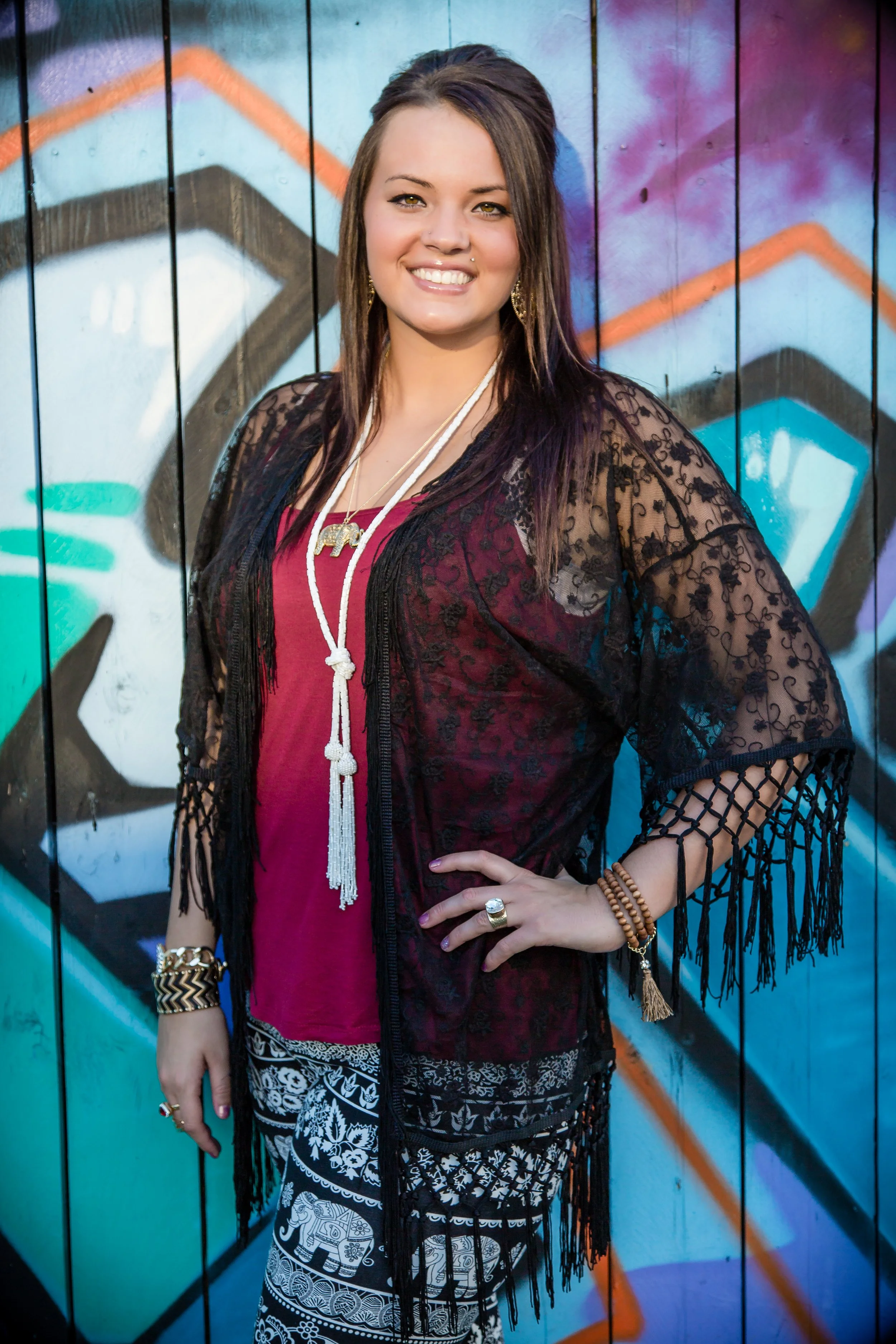 A young woman with brown hair and light skin poses in front of a colorful graffiti wall. She is wearing a black lace cover-up over a maroon top, patterned black and white pants, and various jewelry including a long white beaded necklace, rings, brace