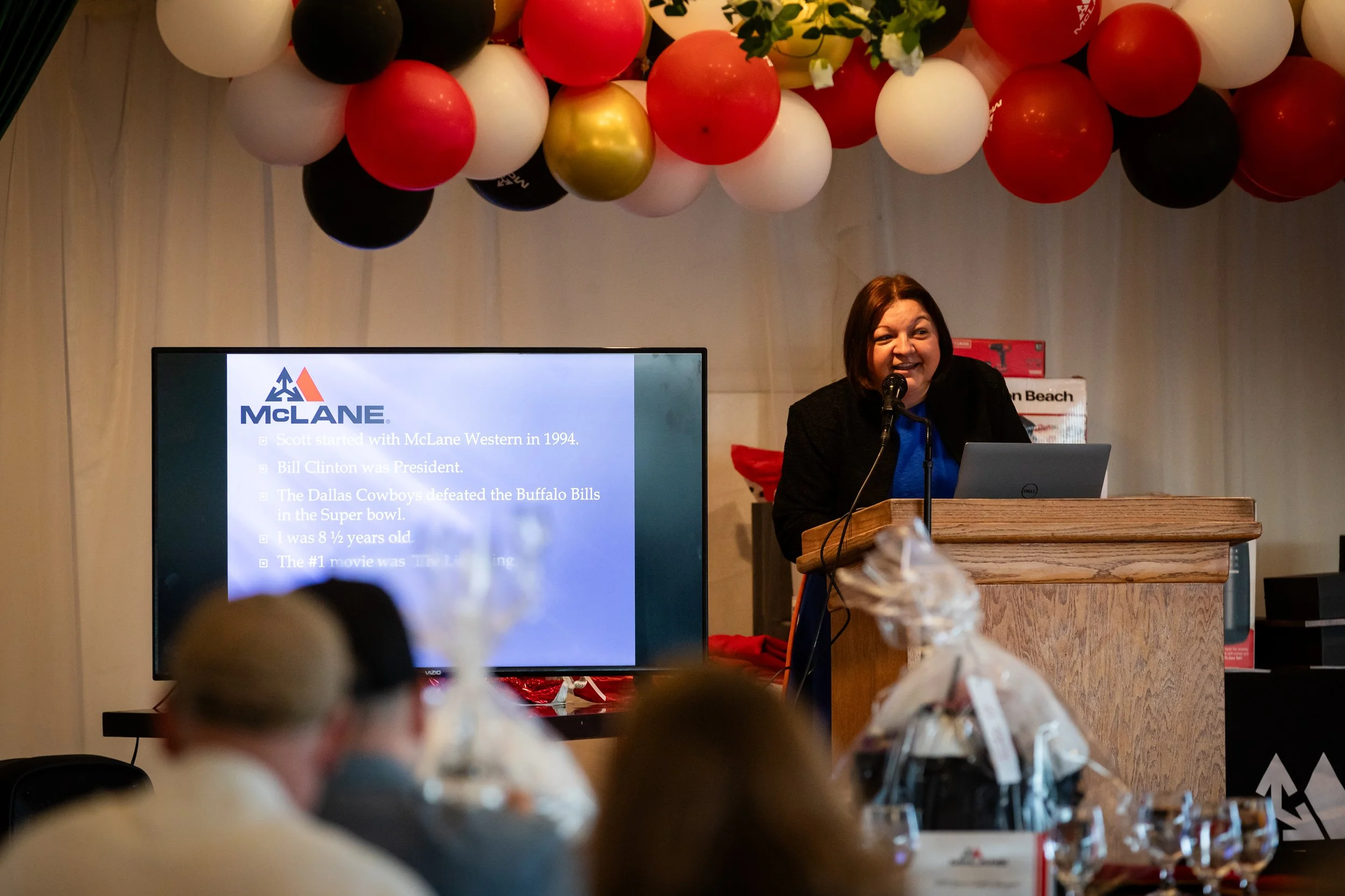 A woman speaking at a presentation with balloons above and a screen displaying information about McLane. Audience members and tables with decorations are in the foreground.