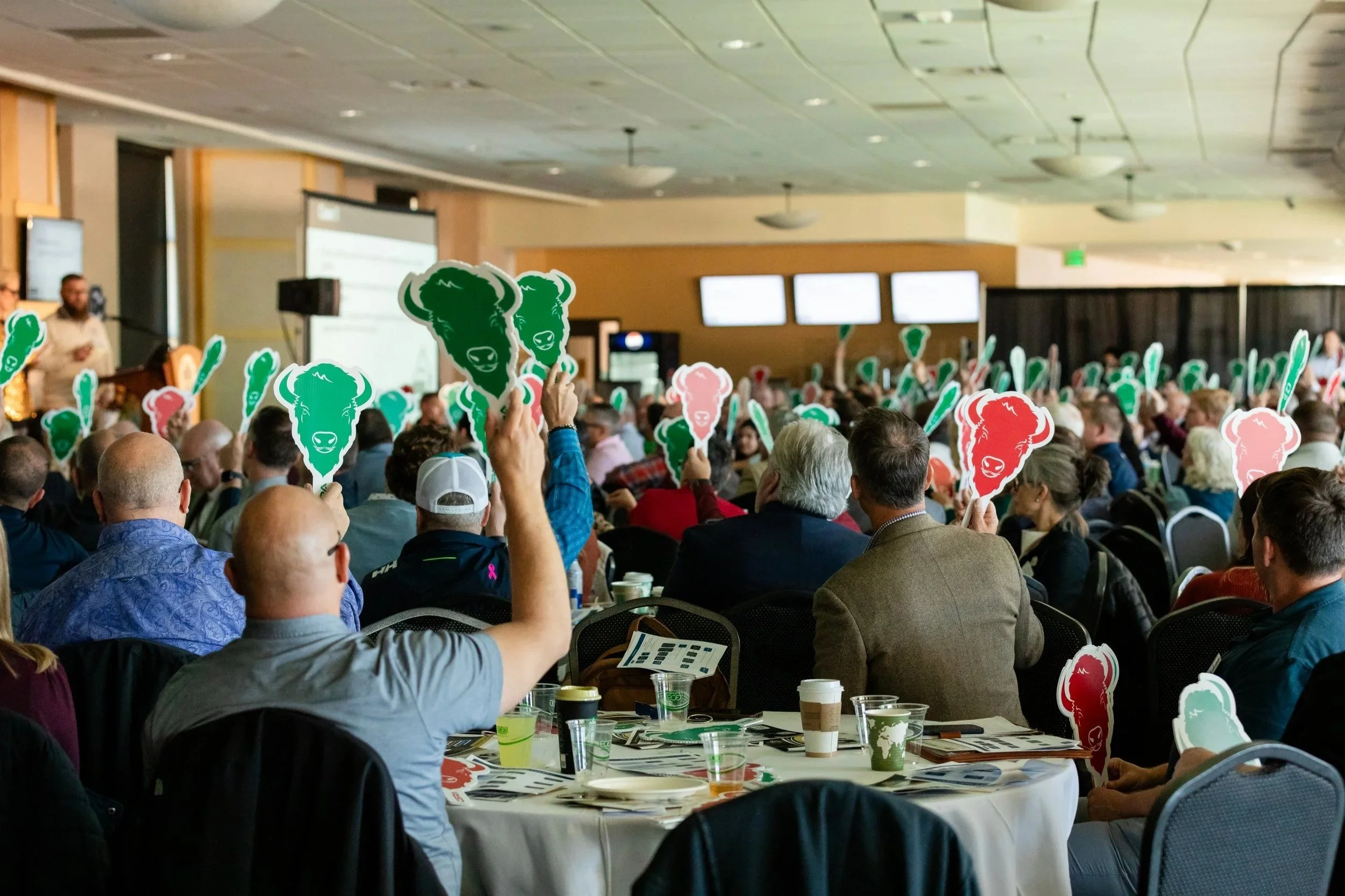 A large indoor presentation or conference with many attendees holding cutouts of a cartoon buffalo head, predominantly in green, red, and pink colors, with some seated at tables and others standing.