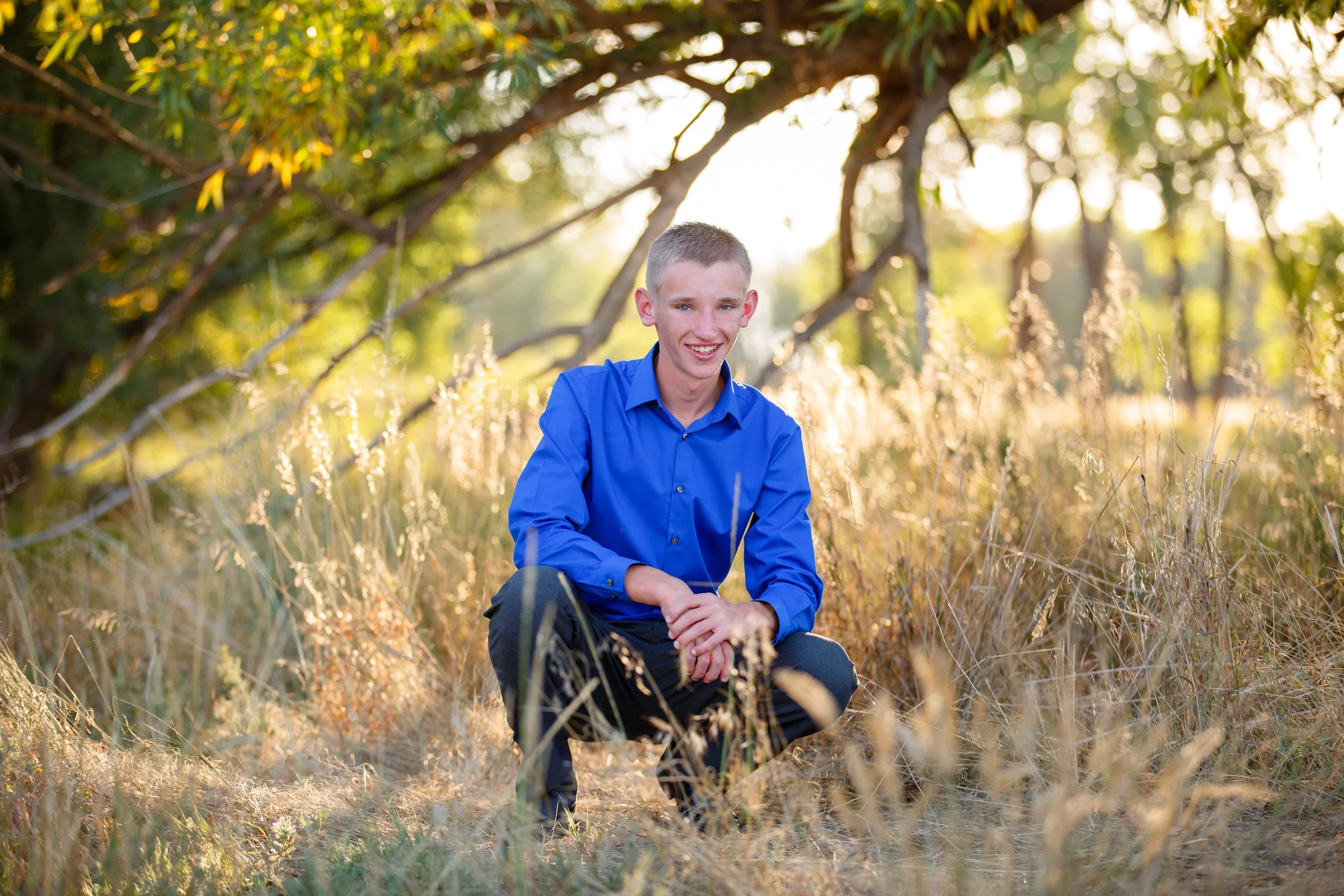 A young man with short blonde hair and blue eyes, wearing a bright blue shirt, smiling while sitting in a grassy, wooded area during golden hour.