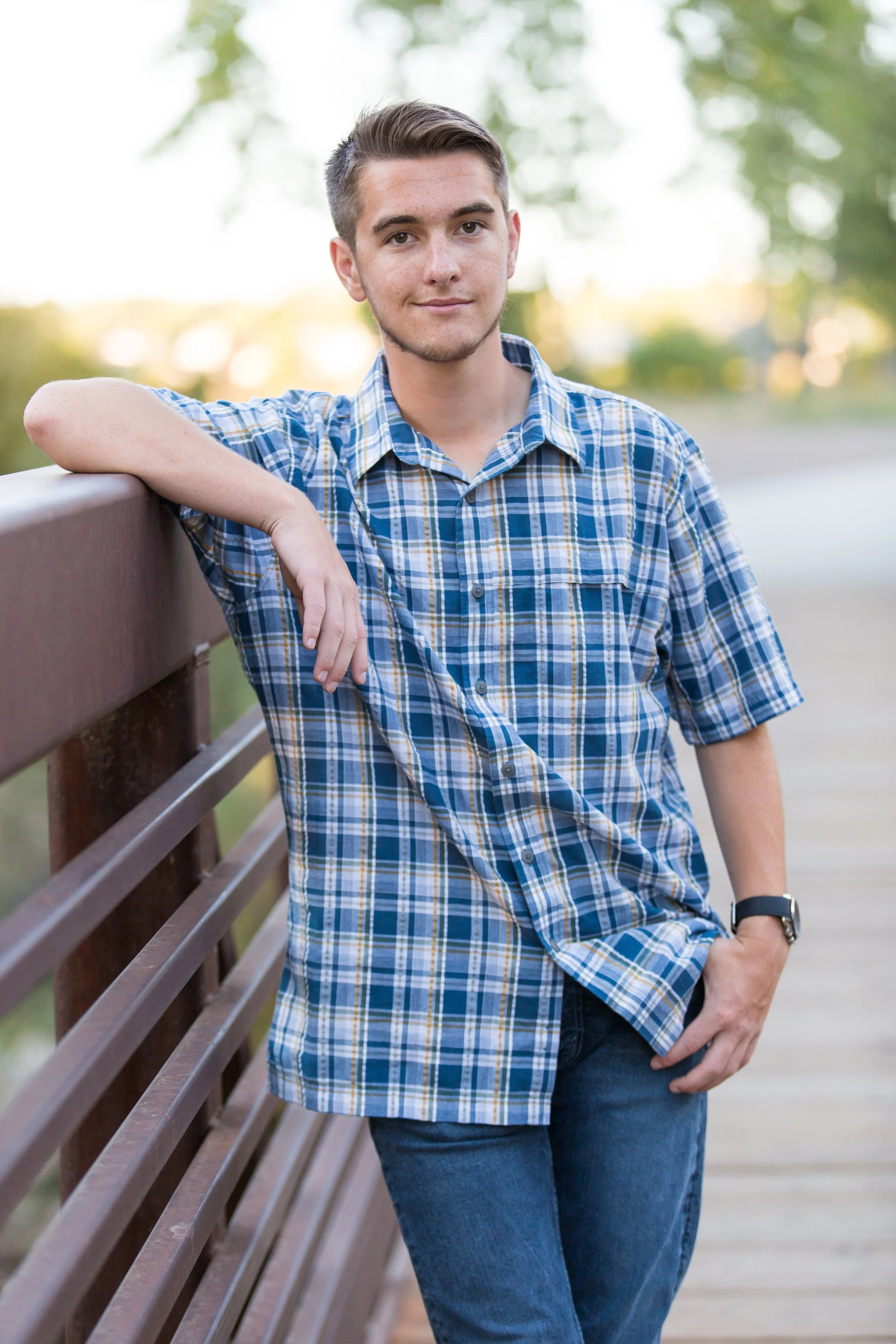 An outdoor high school senior portrait session featuring a young man leaning on a rustic bridge structure on location at Golden Ponds in Longmont, Colorado.
