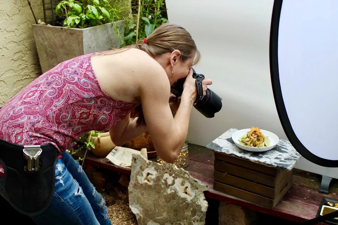 A woman taking a photograph of a plate of food outdoors, with a large light diffuser nearby.