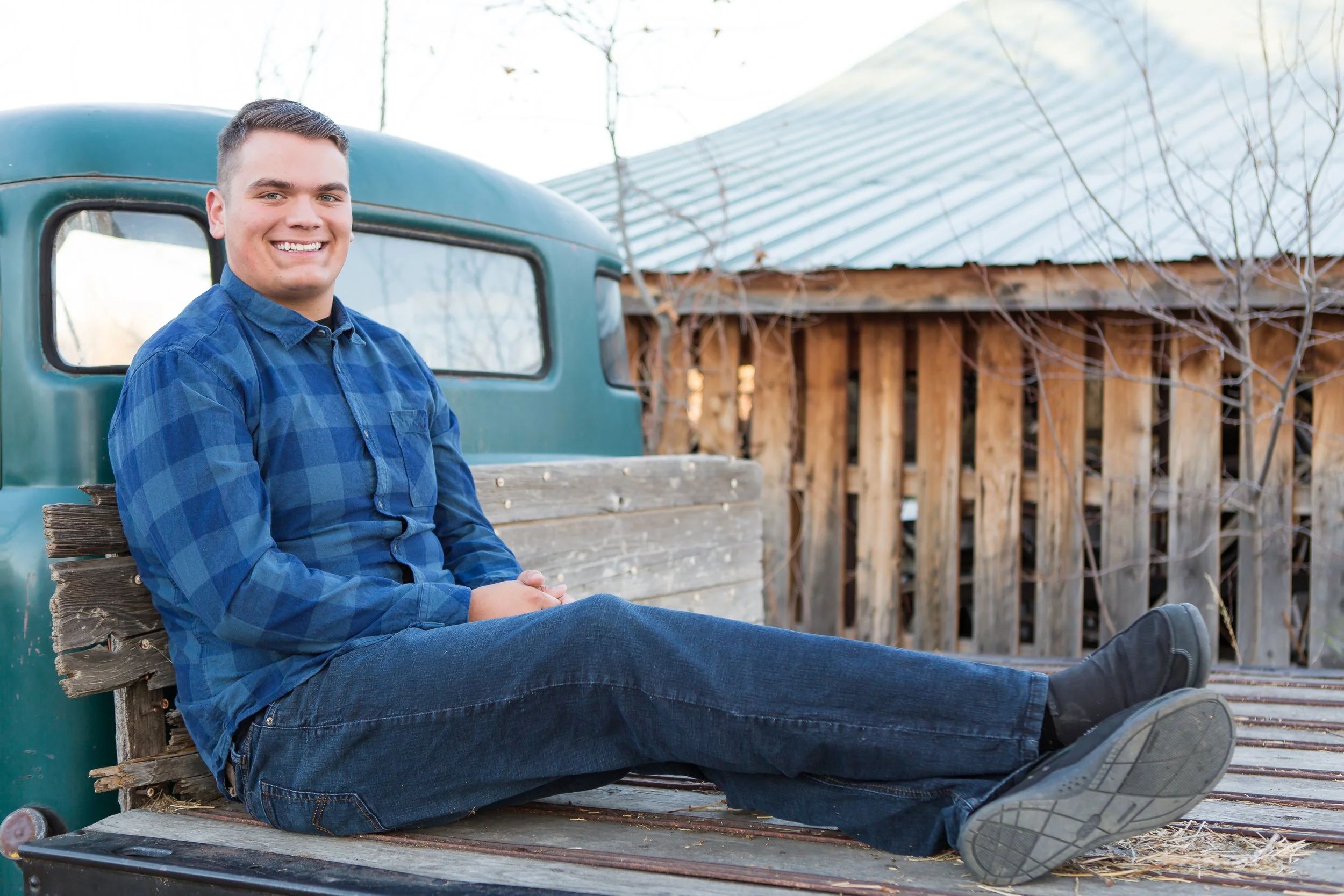 A young man sitting on a wooden bench in front of an old farm truck, smiling at the camera with a barn and leafless trees in the background.