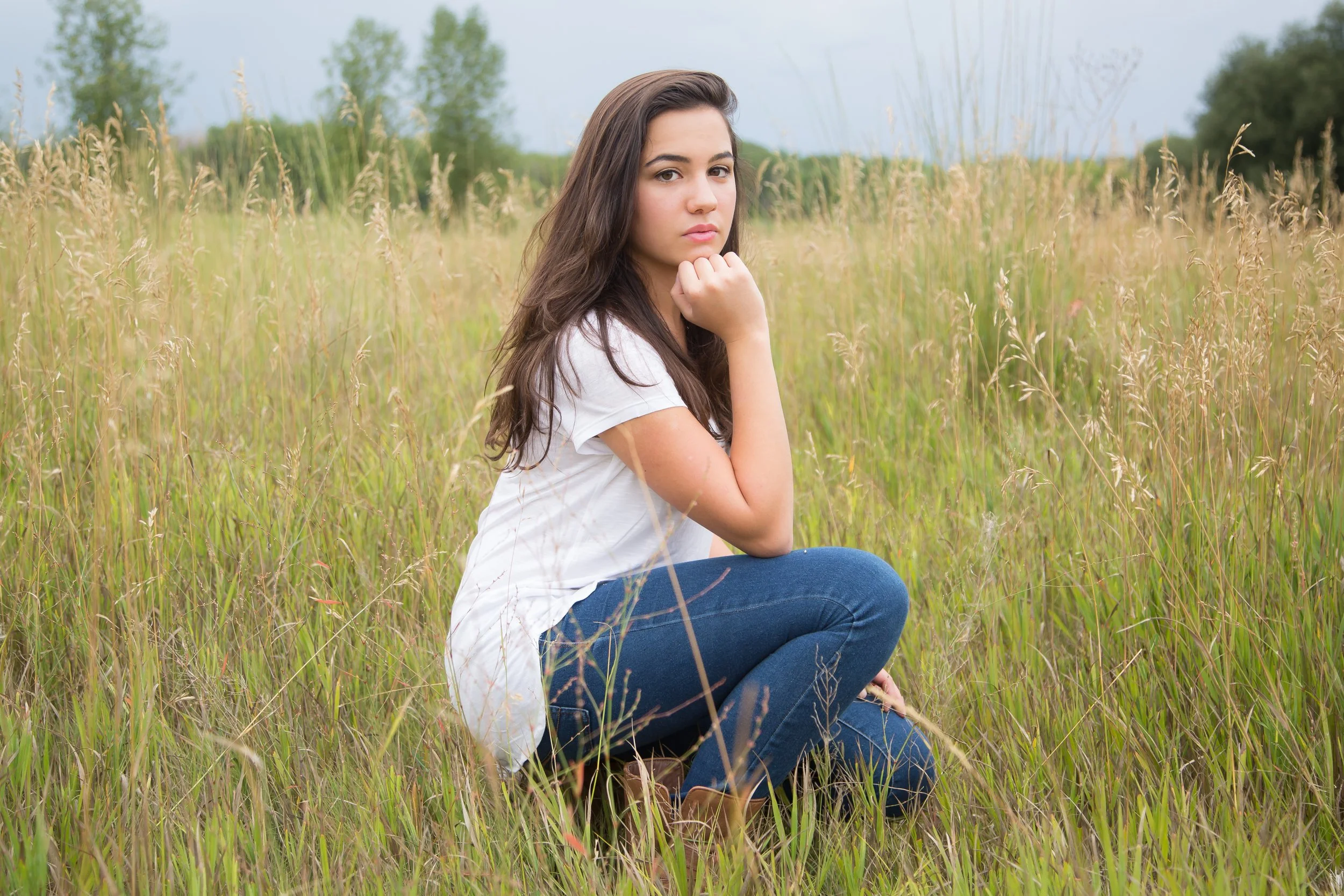 A young woman with long dark hair, sitting in a grassy field with tall wild grass and trees in the background, looking at the camera with a neutral expression.