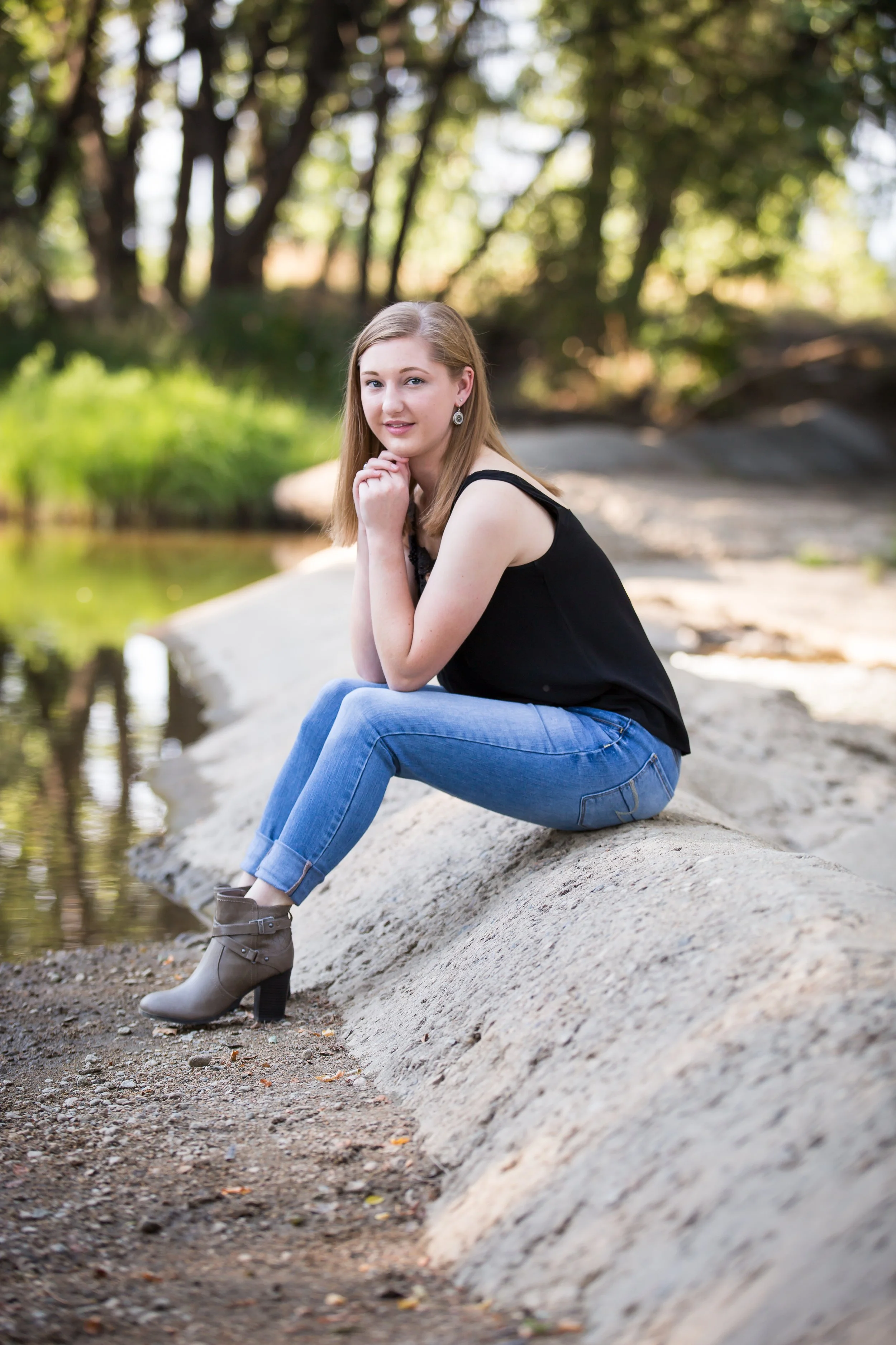 A young woman with long blonde hair, wearing a black tank top, blue jeans, and beige ankle boots, sitting on a large rock near a small pond in a wooded area during daylight, with trees and greenery in the background.