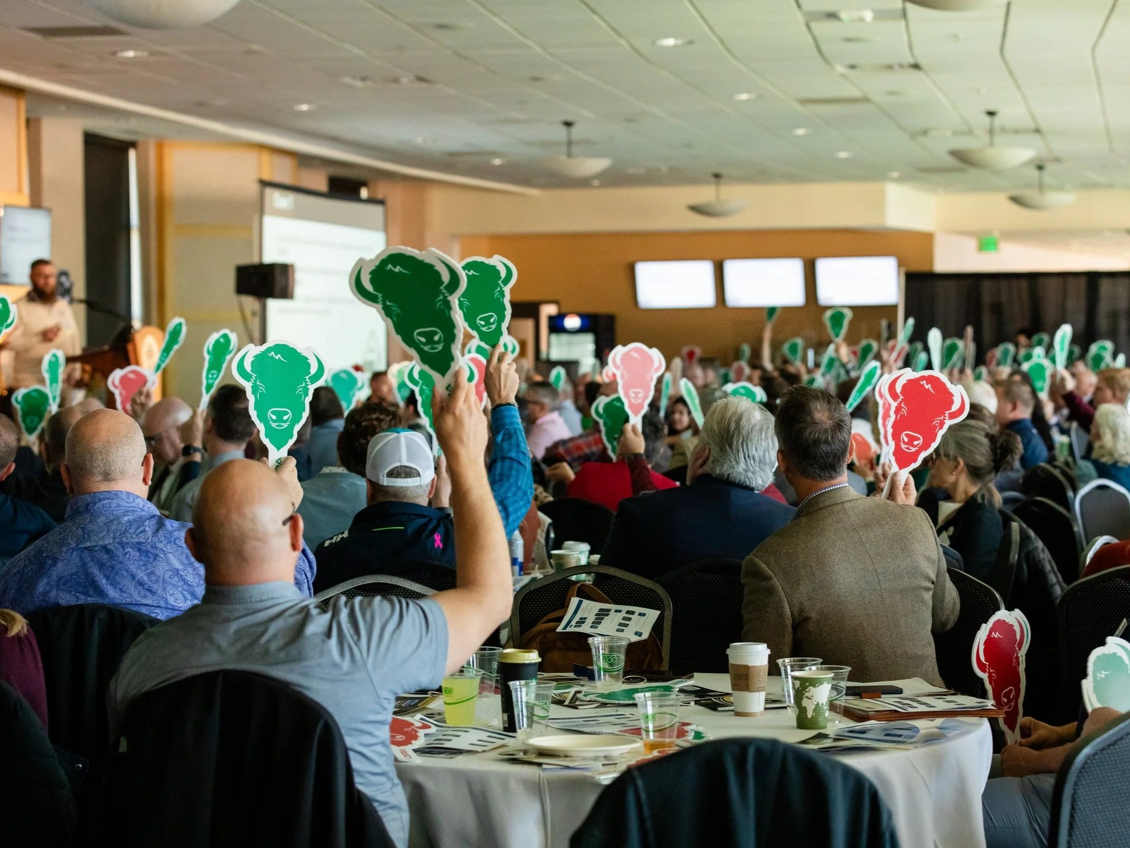Attendees at a conference hold up cutouts of green and red buffalo heads with smiling faces, seated in a large room with multiple screens and a presenter at the front.