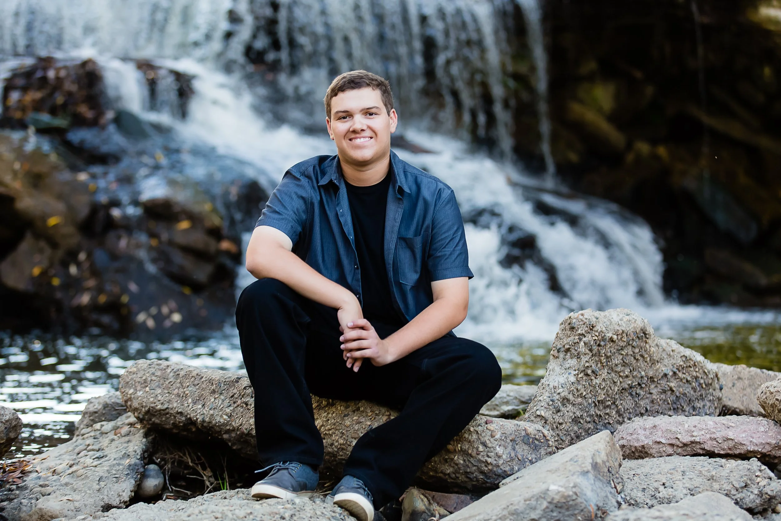 A young man sitting on rocks near a waterfall and a small stream, smiling at the camera.