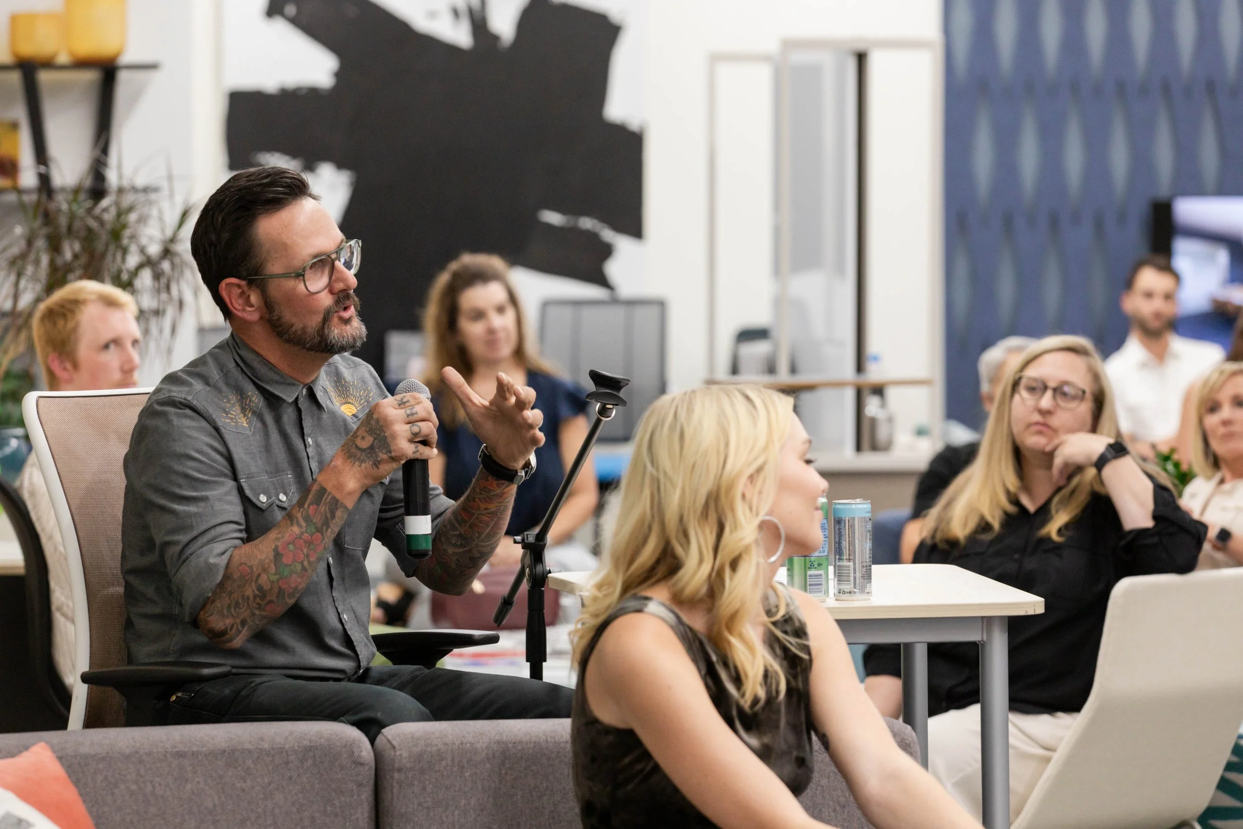 A man with tattoos, glasses, and a beard, holding a microphone, speaking during a meeting or discussion while people listen attentively.