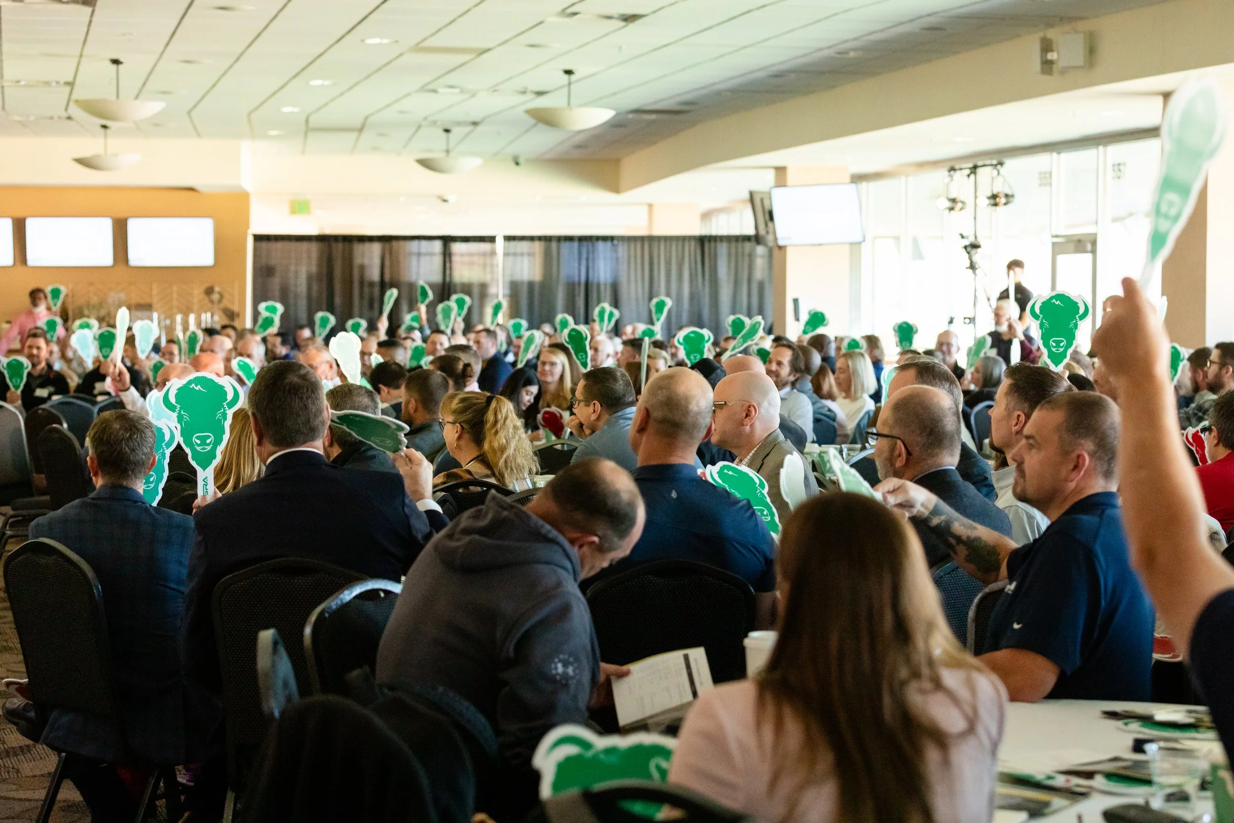 A large indoor conference room filled with a diverse group of people sitting at tables, raising green and white foam hands with a buffalo head and a speech bubble. People are engaging and looking towards the front, with large windows letting in natur