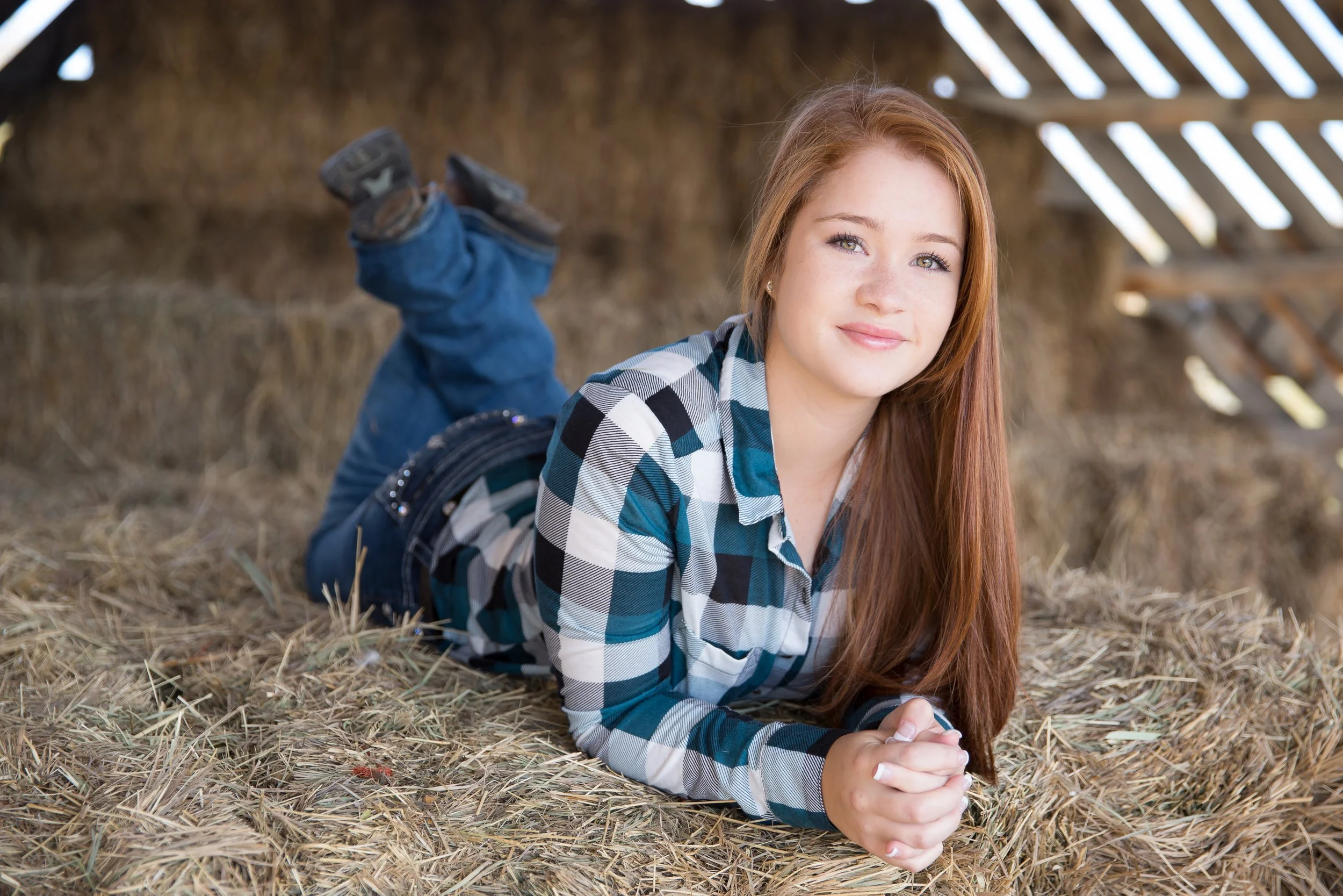 A rustic high school senior portrait session featuring a smiling girl lying on a bed of hay inside the historic hay loft at Lone Hawk Farm in Longmont, Colorado.