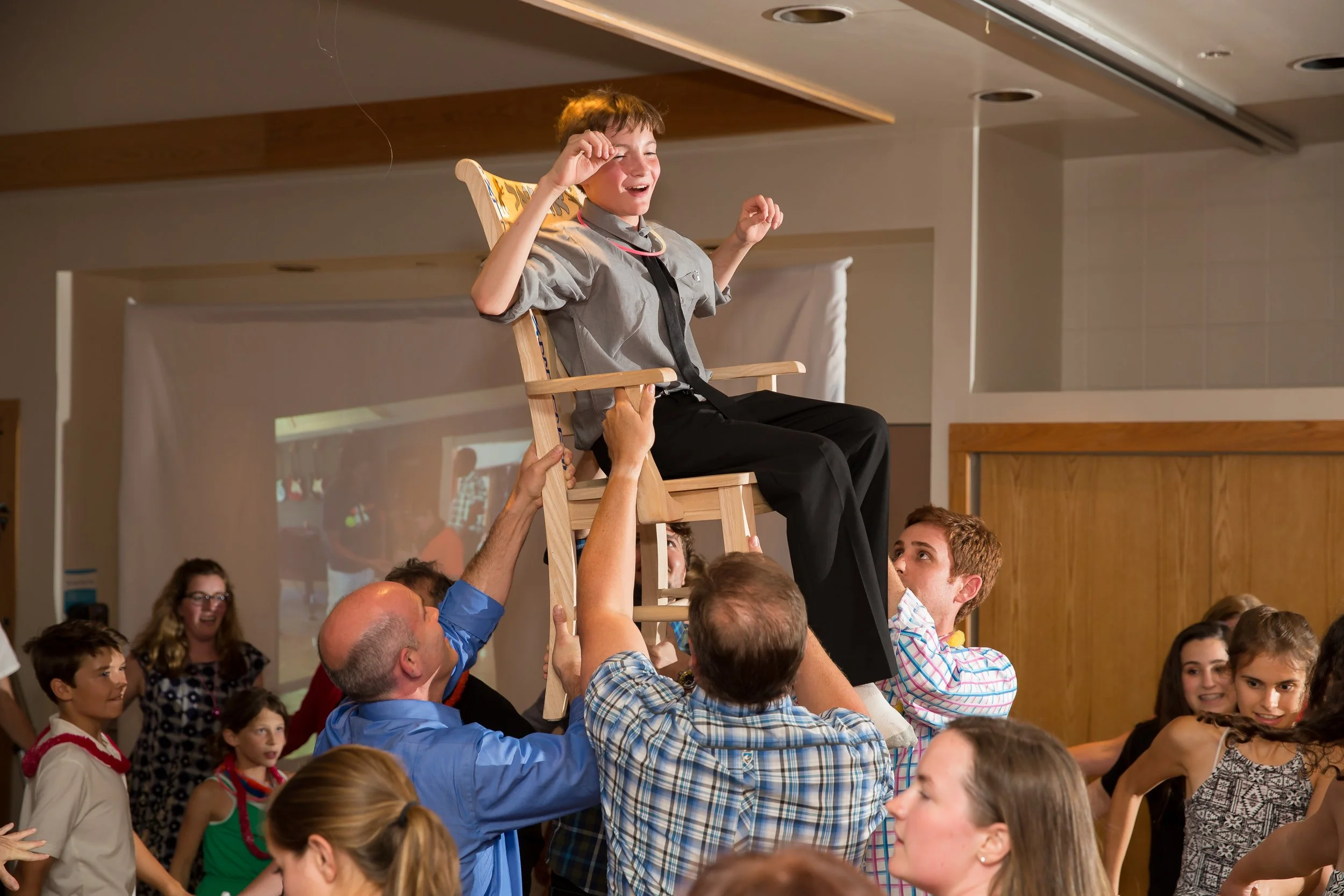 A high-energy documentary style event photograph of a boy being raised on a wooden chair during the traditional Hora dance at a Bar Mitzvah celebration, by Lisa Doane Photography.