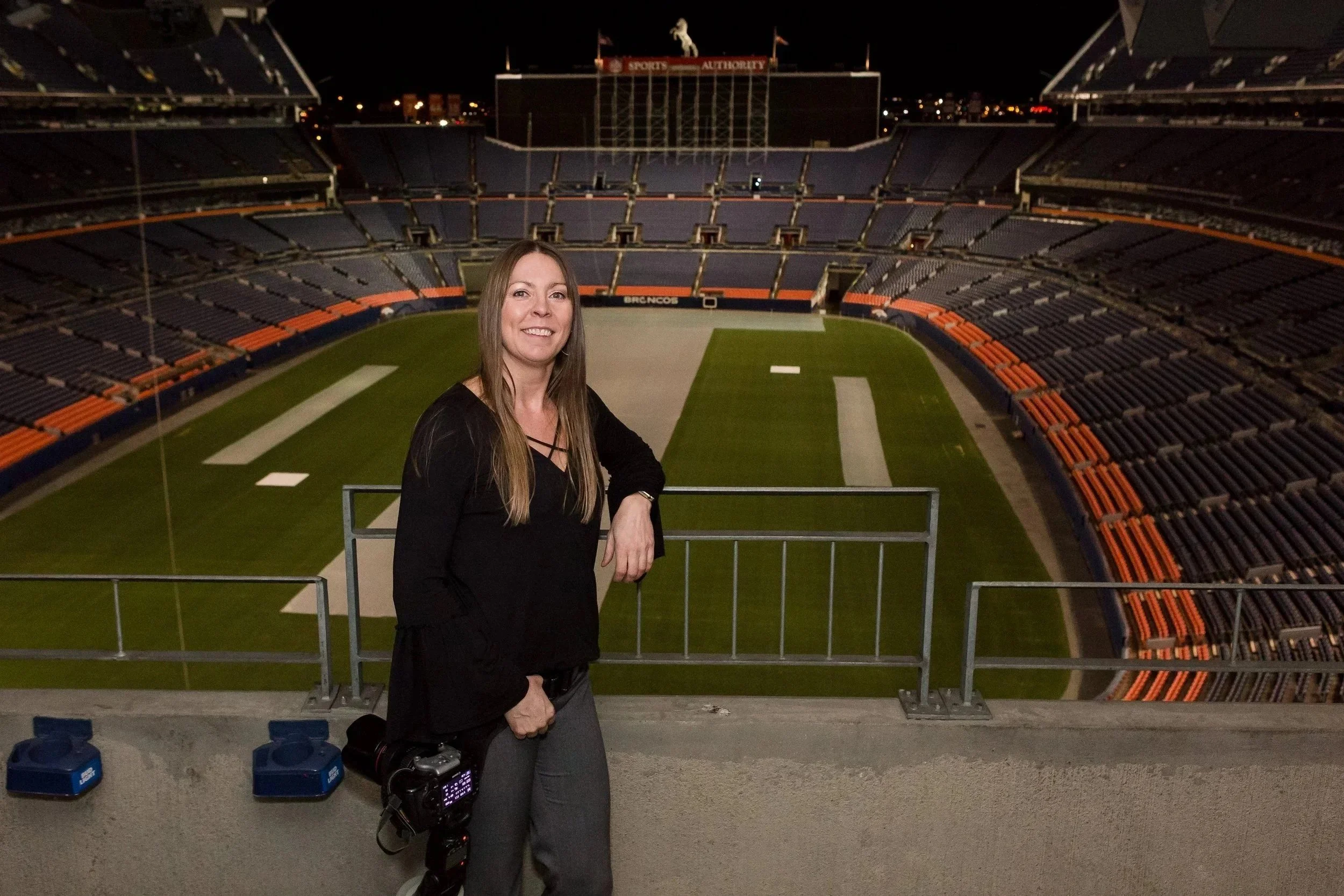 A woman standing in a sports stadium at night, smiling at the camera. She is near the railing on the field with empty seats and a large goalpost visible in the background.