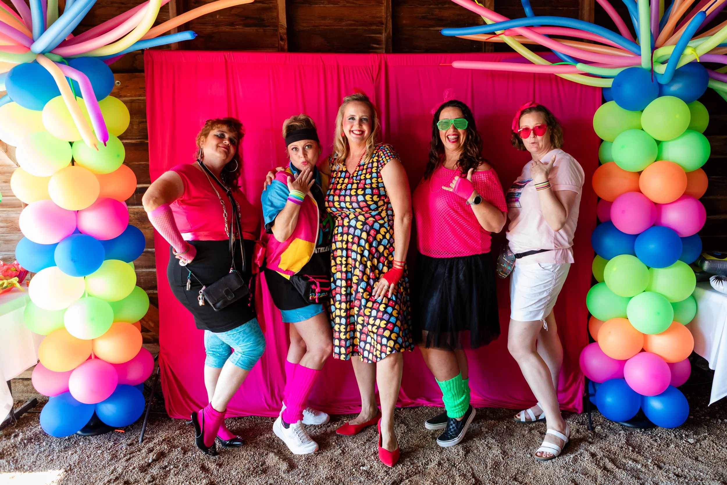 A fun and colorful group portrait of friends posing under a vibrant balloon arch at an 80s themed birthday party photo booth, captured by Lisa Doane Photography.