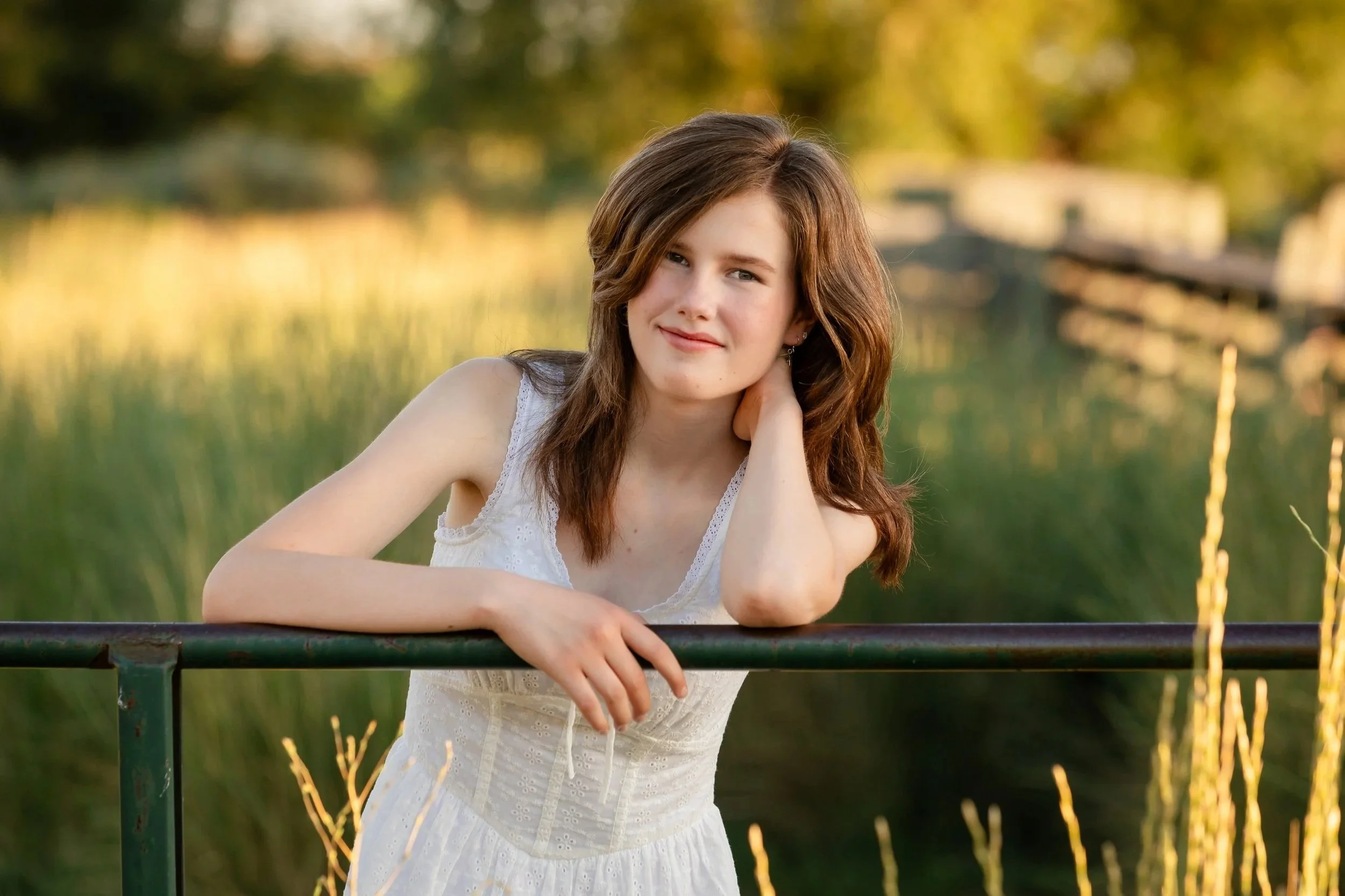 A young woman with wavy brown hair and fair skin leaning on a metal railing outdoors in a park or garden during sunset, wearing a white sleeveless dress with lace details, with a calm expression on her face.