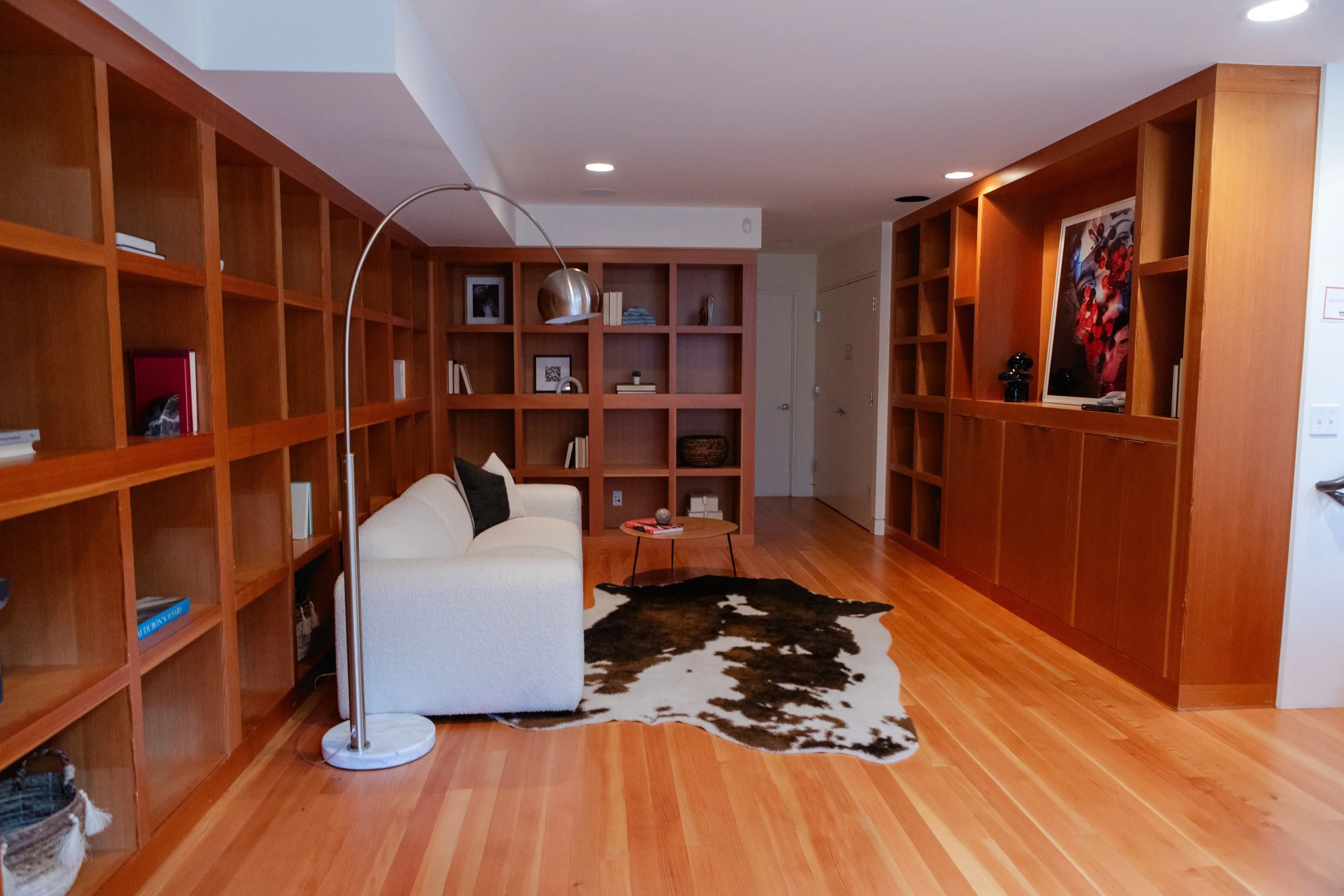 Living room with wooden built-in bookshelves, a white sofa with black pillows, a cowhide rug, a small round coffee table, and a modern floor lamp. The shelves contain books, framed photos, and decorative items. The room has wooden floors and white wa