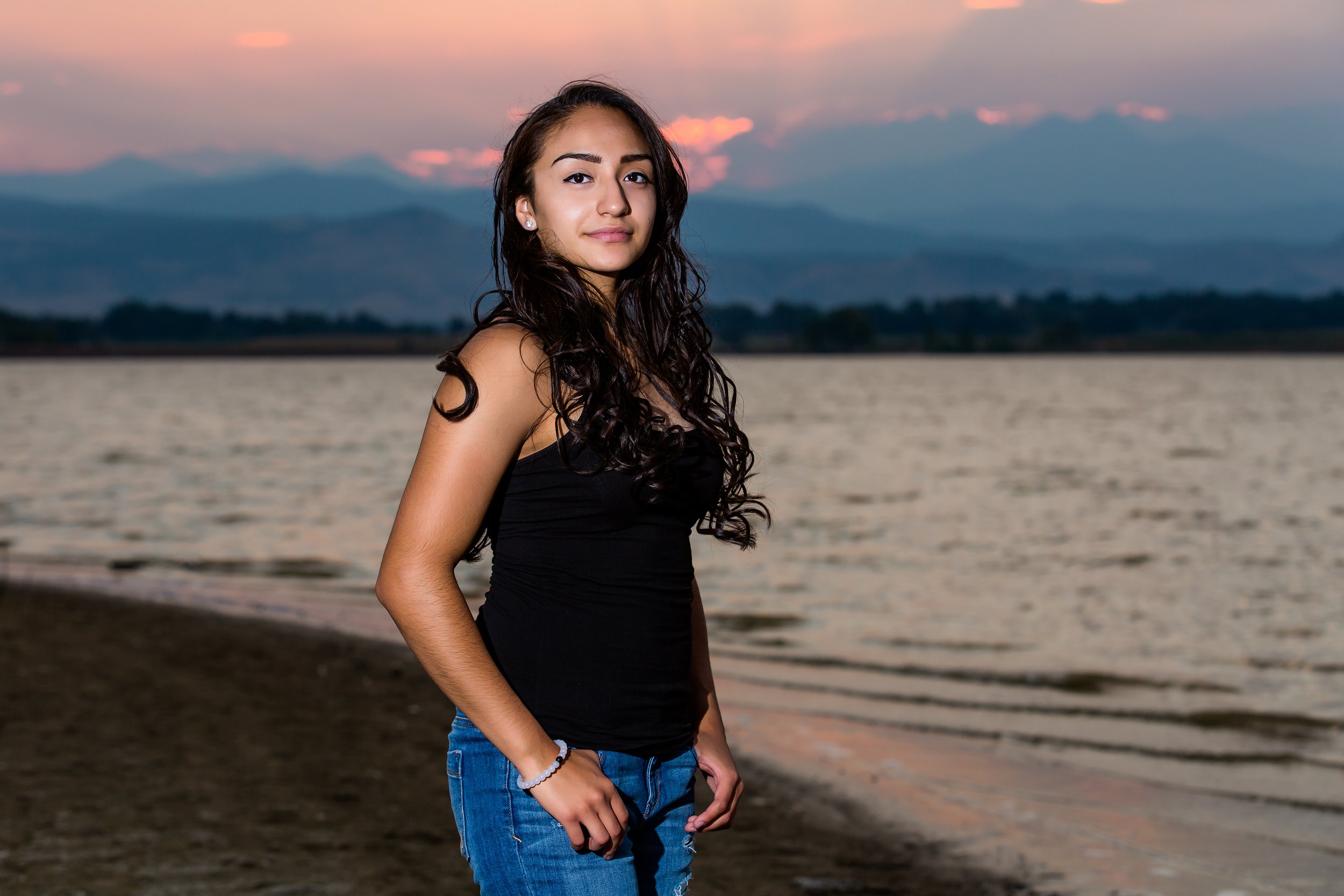 A high school senior portrait session of a girl posing along the shoreline on the southwest side of McIntosh Lake in Longmont, Colorado, during a scenic sunset with mountains in the background.