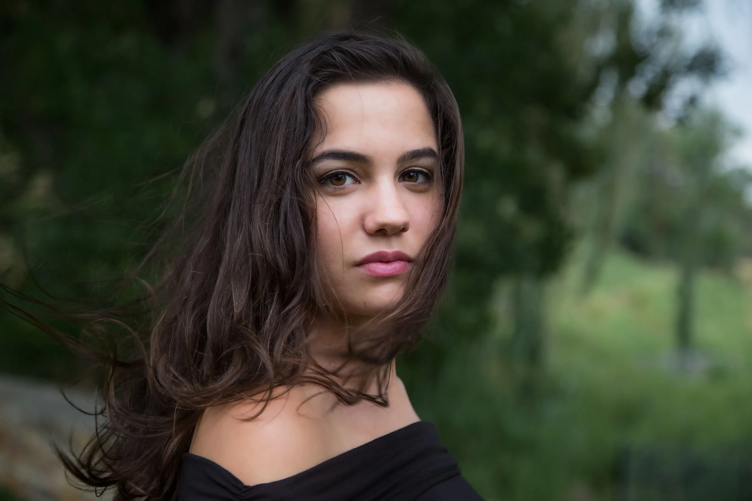 A young woman with long, wavy brown hair and brown eyes, wearing a black off-the-shoulder top, standing outdoors with blurred green foliage in the background.