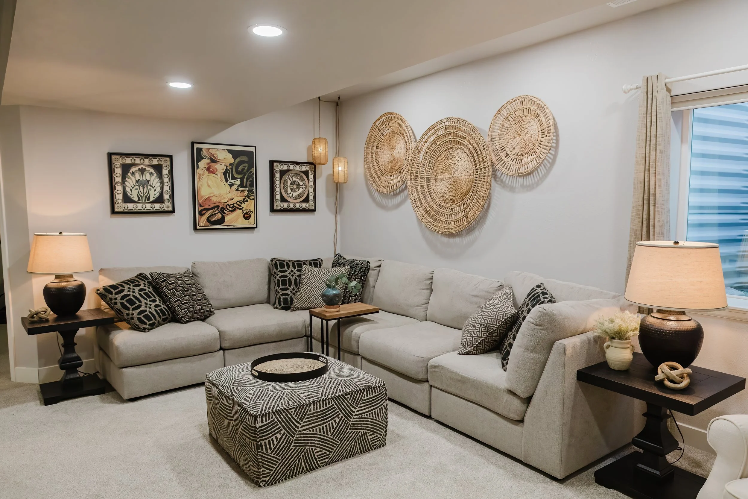 Living room with white sectional sofa, patterned pillows, black side tables with lamps, wall art, and woven wall decor, next to a window with beige curtains.
