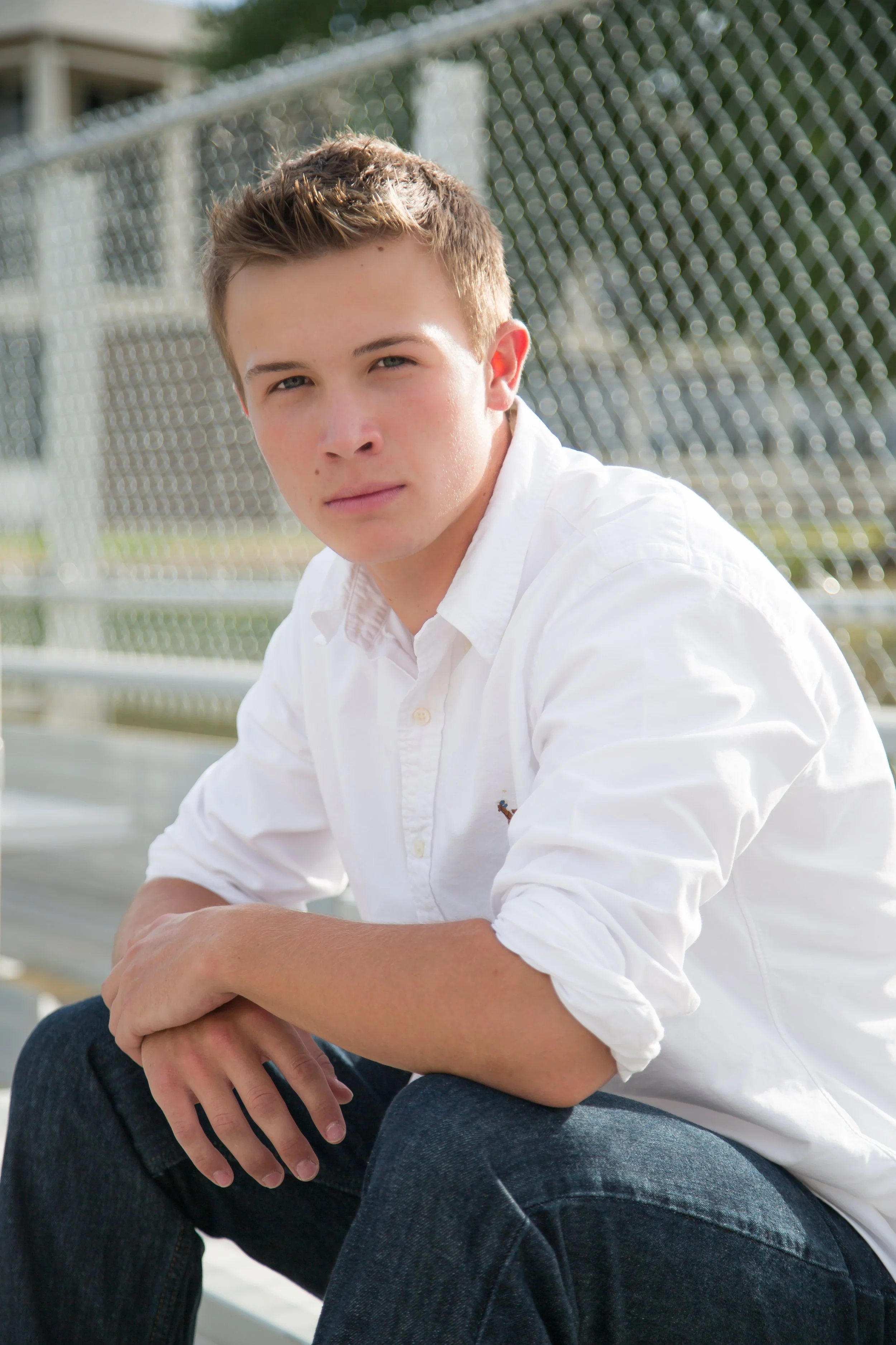 A young man with short blonde hair wearing a white collared shirt sitting outdoors near a chain-link fence.