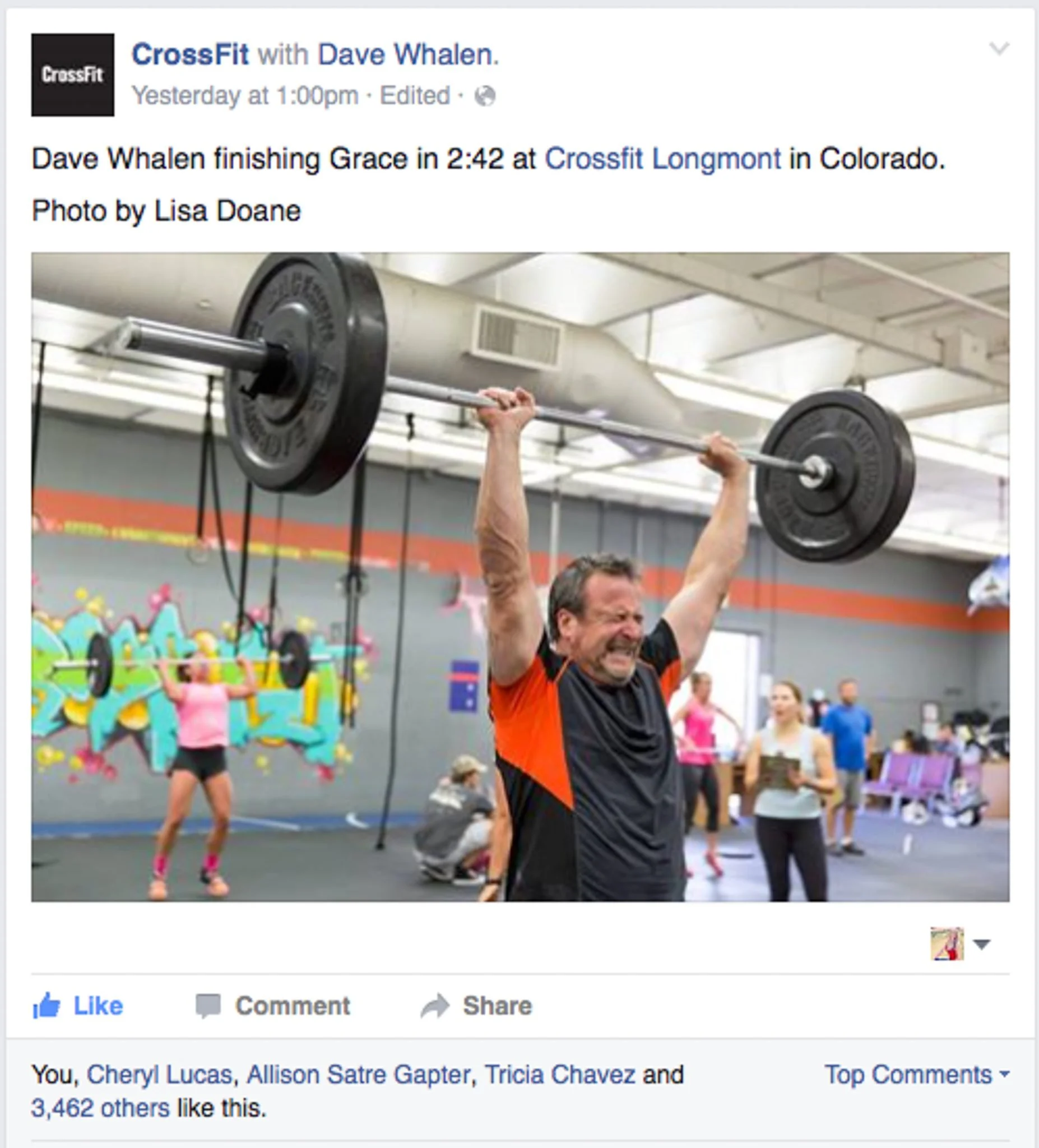 A man lifting barbell weights in a gym. The background shows a woman doing exercises and colorful decorations on the wall.