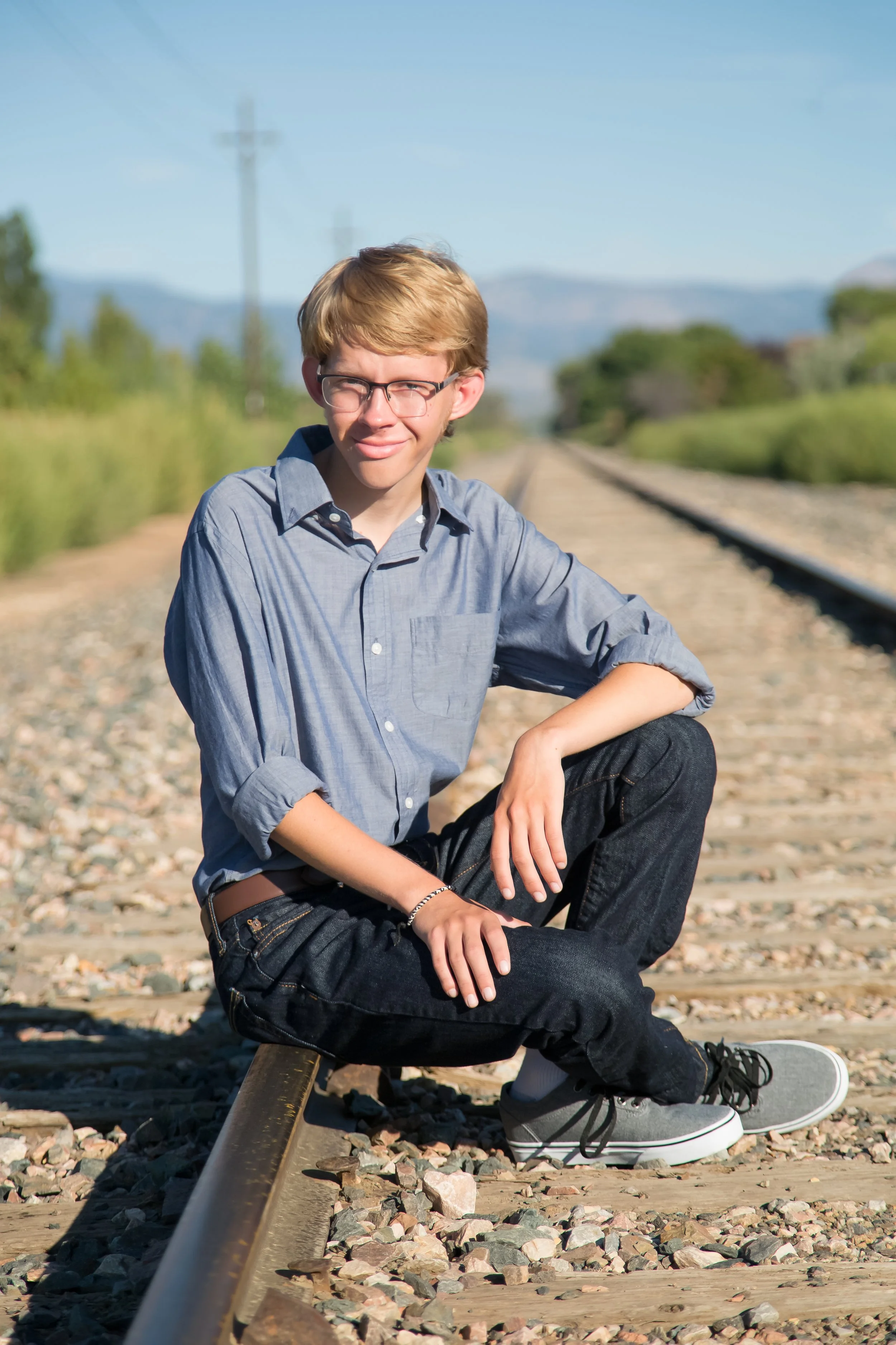 A rustic high school senior portrait session featuring a young man sitting on the railway tracks with a scenic background of trees and mountains at Golden Ponds in Longmont, Colorado.