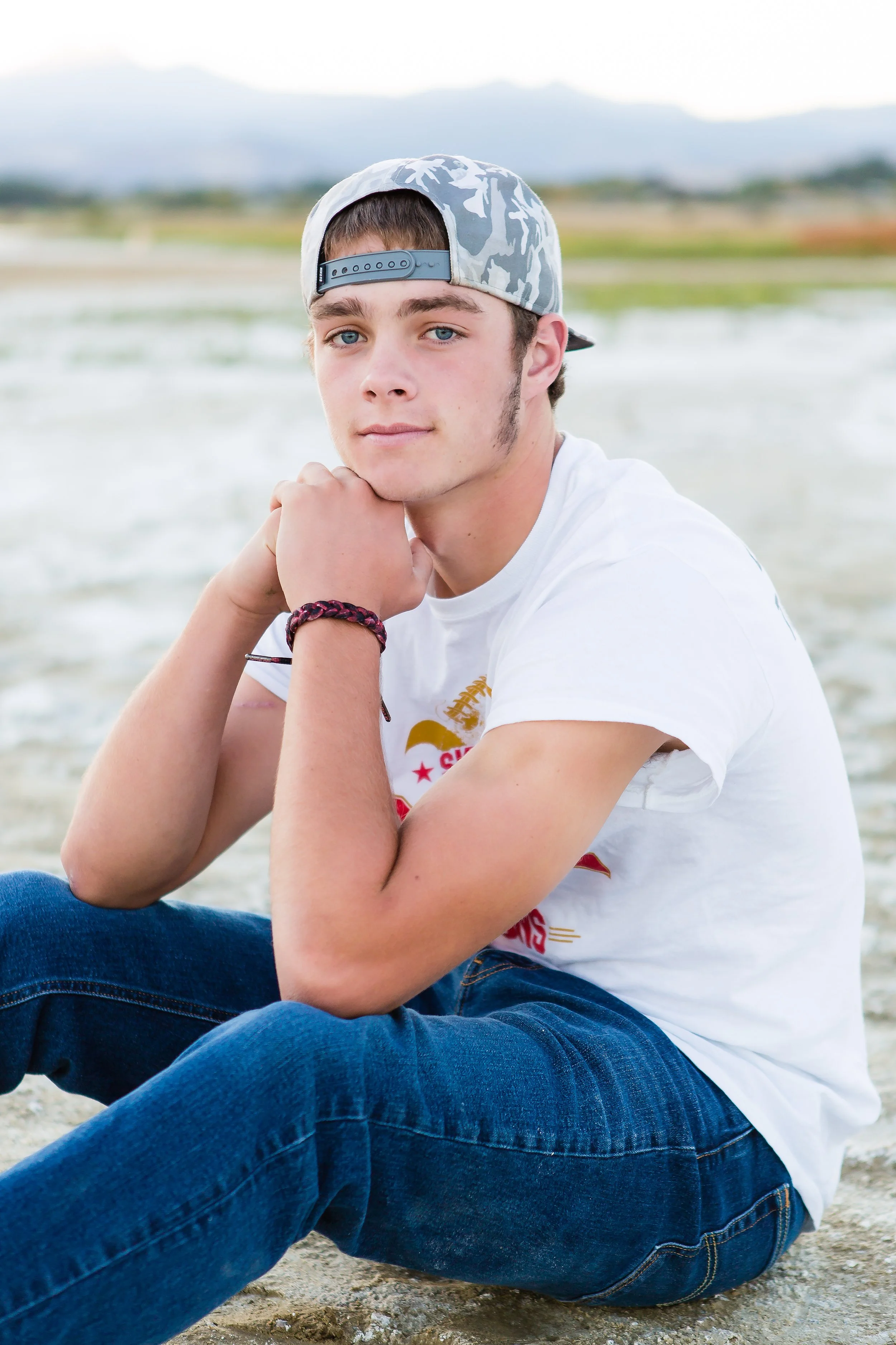 A young man with blue eyes and light brown hair sitting on a sandy beach, wearing a camouflage cap backward, a white t-shirt, and blue jeans, with mountains in the background.