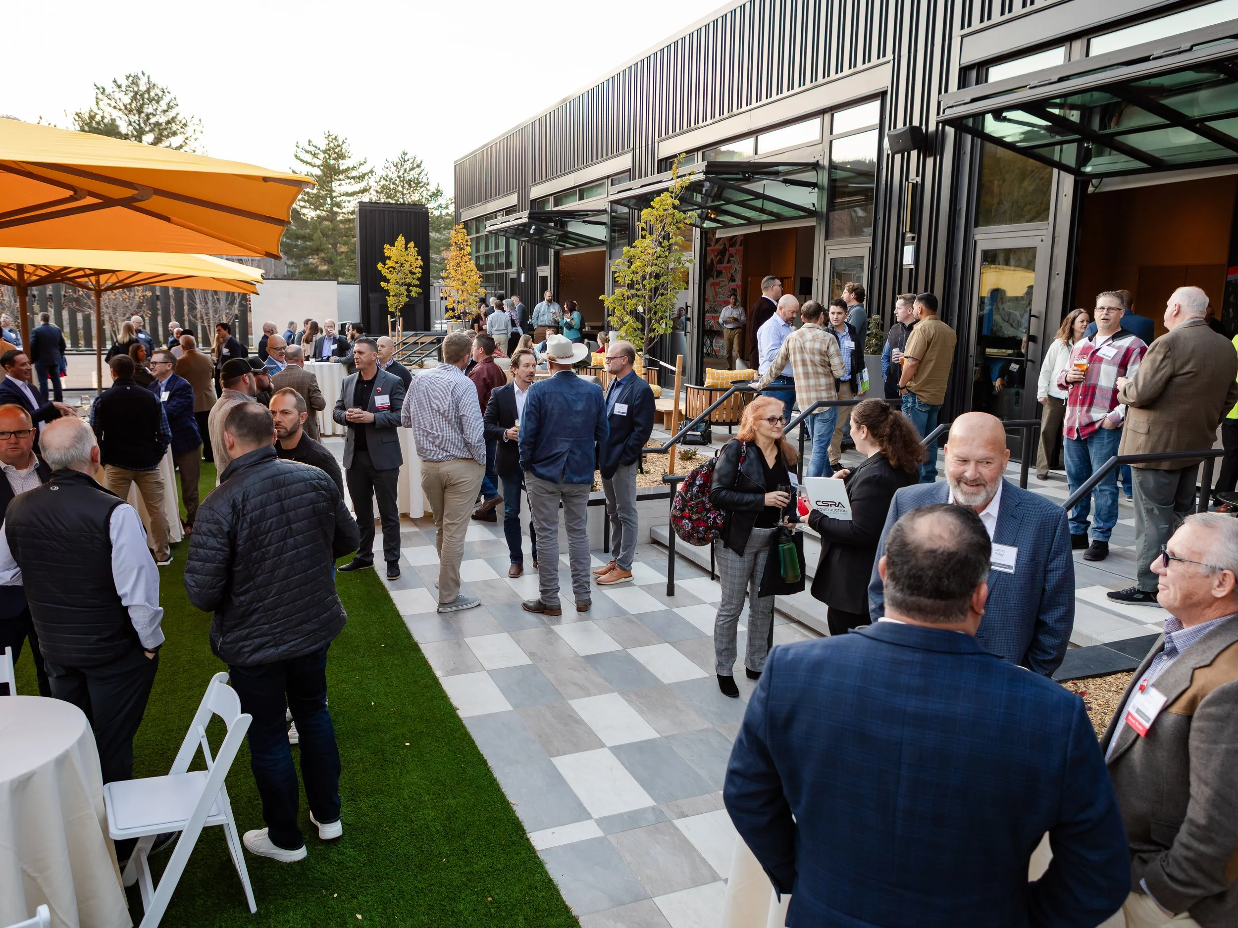 Outdoor networking event with people socializing on a patio with black building exterior, yellow umbrellas, and some greenery.