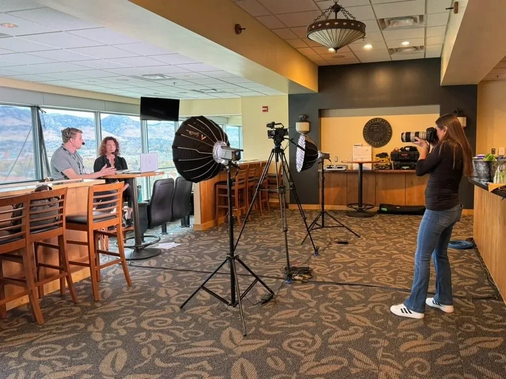 A woman filming two people during an interview in a room with large windows, professional lighting, and camera equipment.