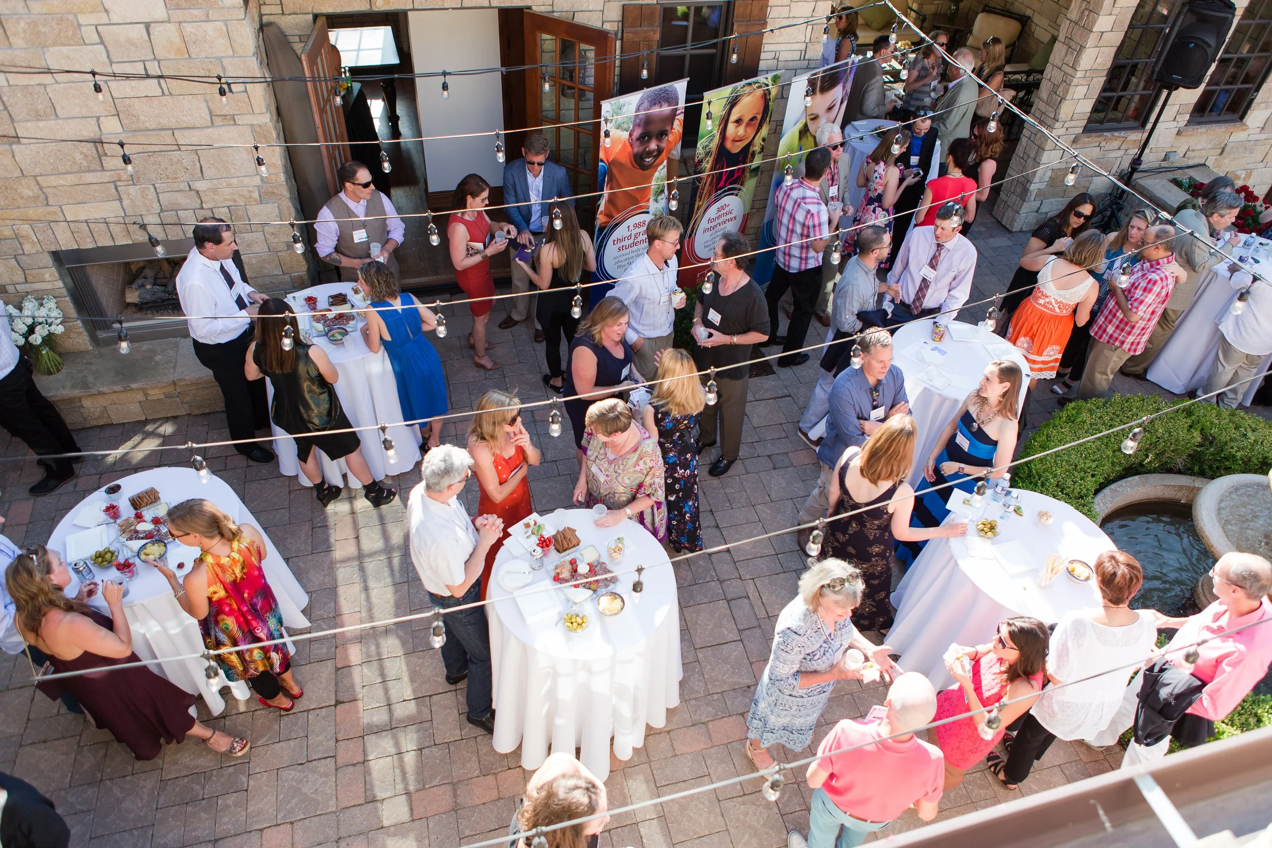 An outdoor social gathering with people mingling around high-top and round tables on a stone patio, decorated with string lights and large portrait banners, at a sunny daytime event.