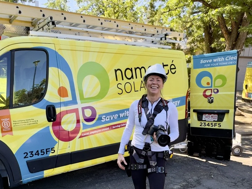 A smiling woman in safety gear, including a white helmet and harness, standing in front of a colorful company branded vehicle and equipment for solar energy installation.