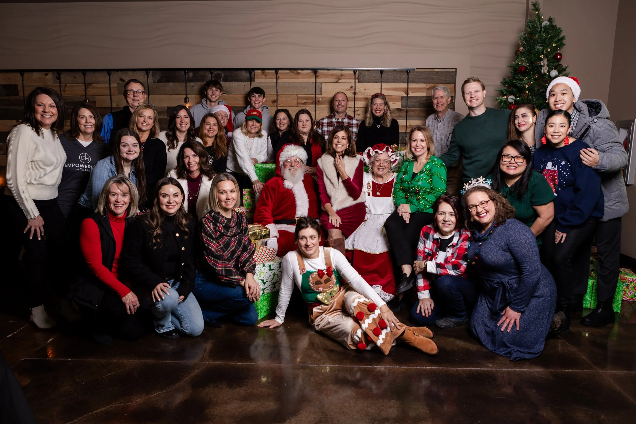 A large group of smiling people, including Santa Claus and Mrs. Claus, gathered together for a Christmas celebration. They are in a decorated indoor space with a Christmas tree and wrapped presents. Many are wearing holiday-themed clothing and access