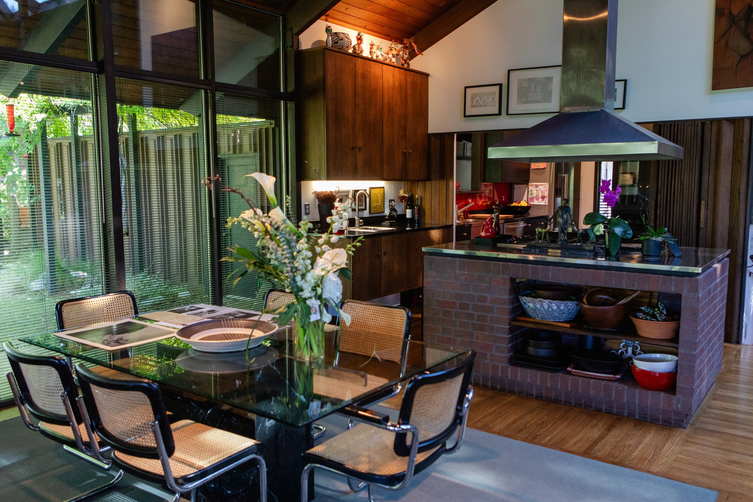 A cozy kitchen and dining area with a glass-topped table and six chairs, flowers in a vase on the table, a kitchen with dark wooden cabinets, and a modern stainless steel range hood over a brick kitchen island, with framed artwork on the walls and de