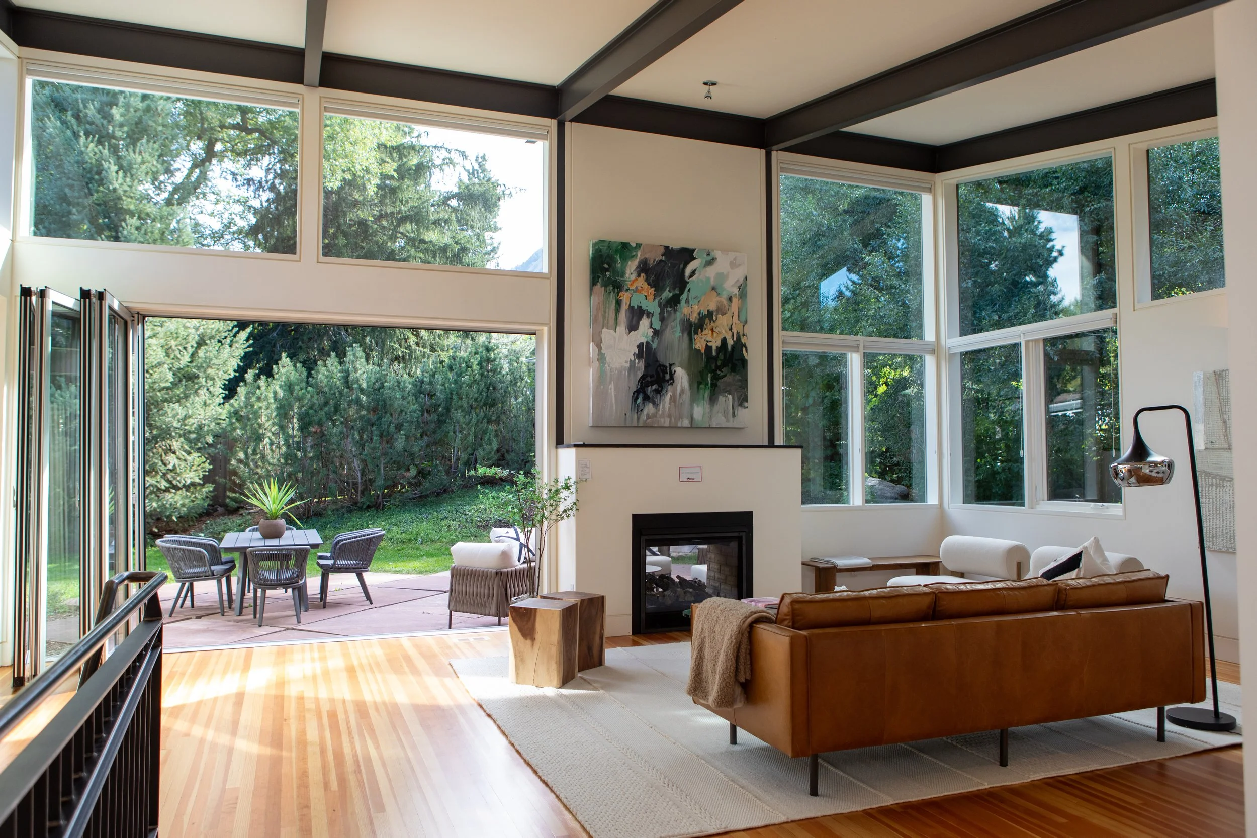 Living room with large windows showing a view of trees and outdoor patio with table and chairs.