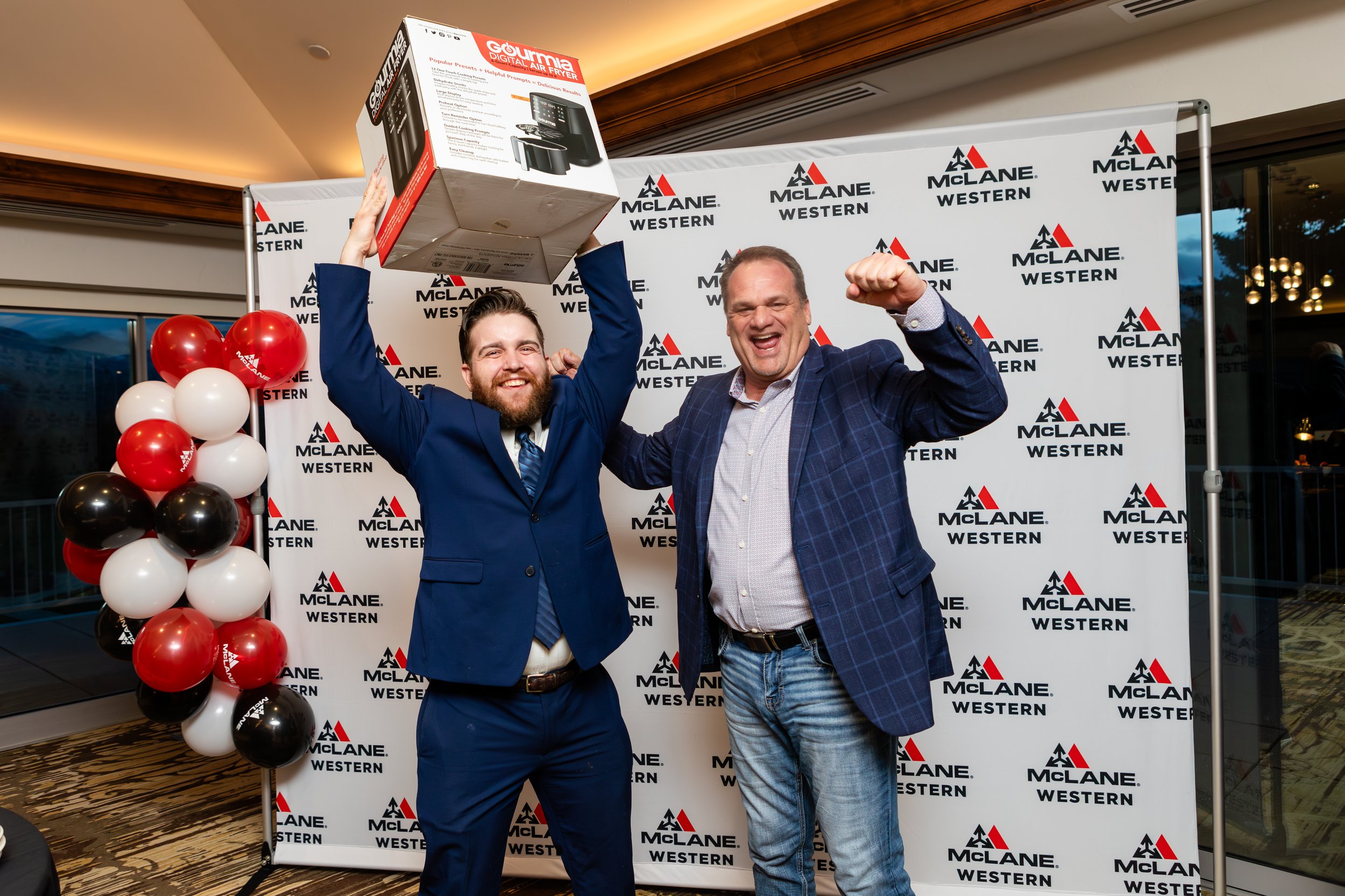 Two men celebrating at an event with a branded McLANE Western backdrop, one holding a box above his head, and balloons in the foreground.