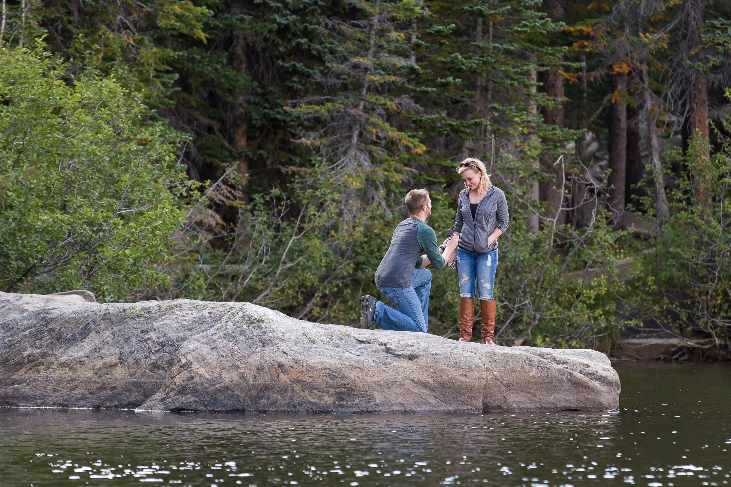 Surprise marriage proposal photography at Bear Lake in Rocky Mountain National Park featuring a man on one knee on a large boulder over the water.
