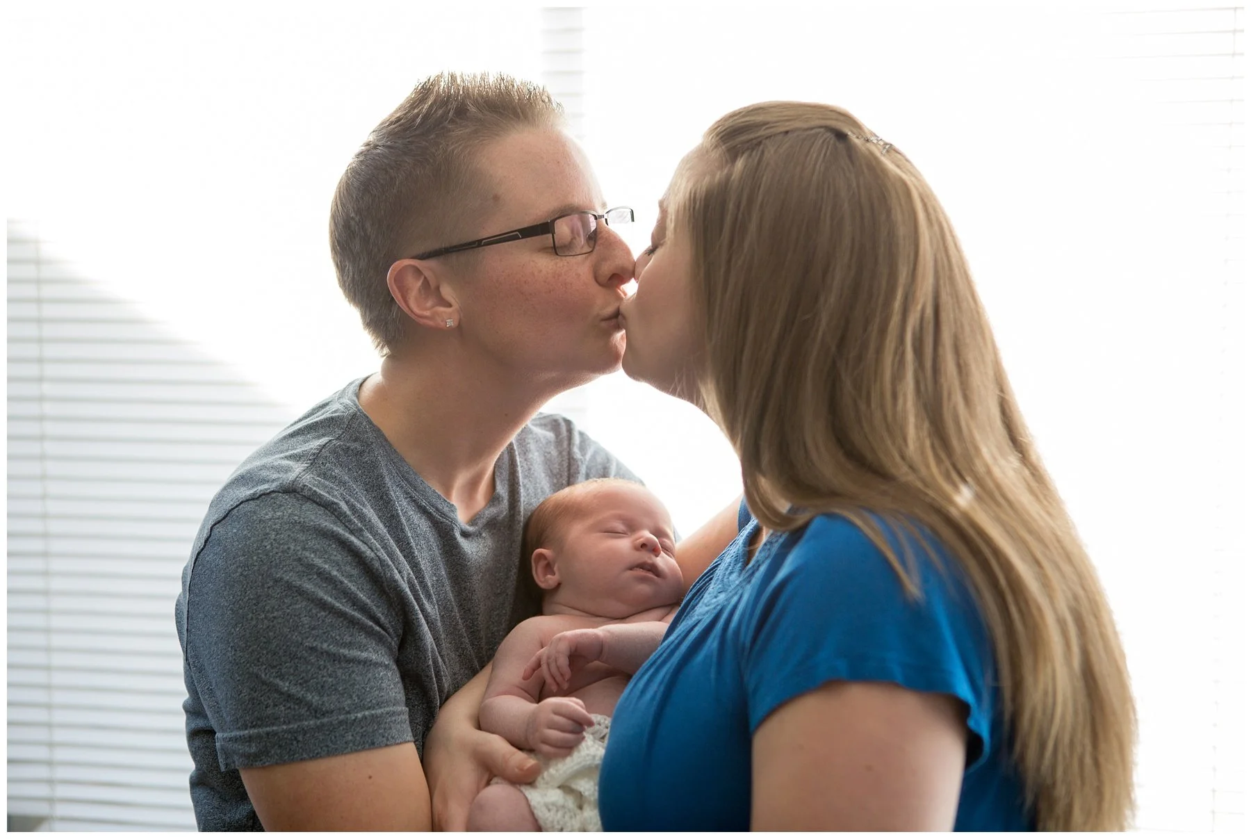 A man and a woman kiss while holding a sleeping baby in their arms in front of a bright window with blinds.