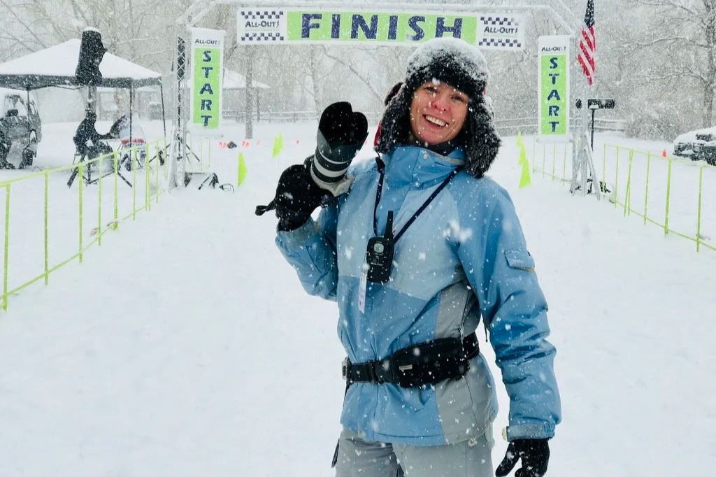 A woman in winter gear holding a camera, smiling at the finish line of a snow race.