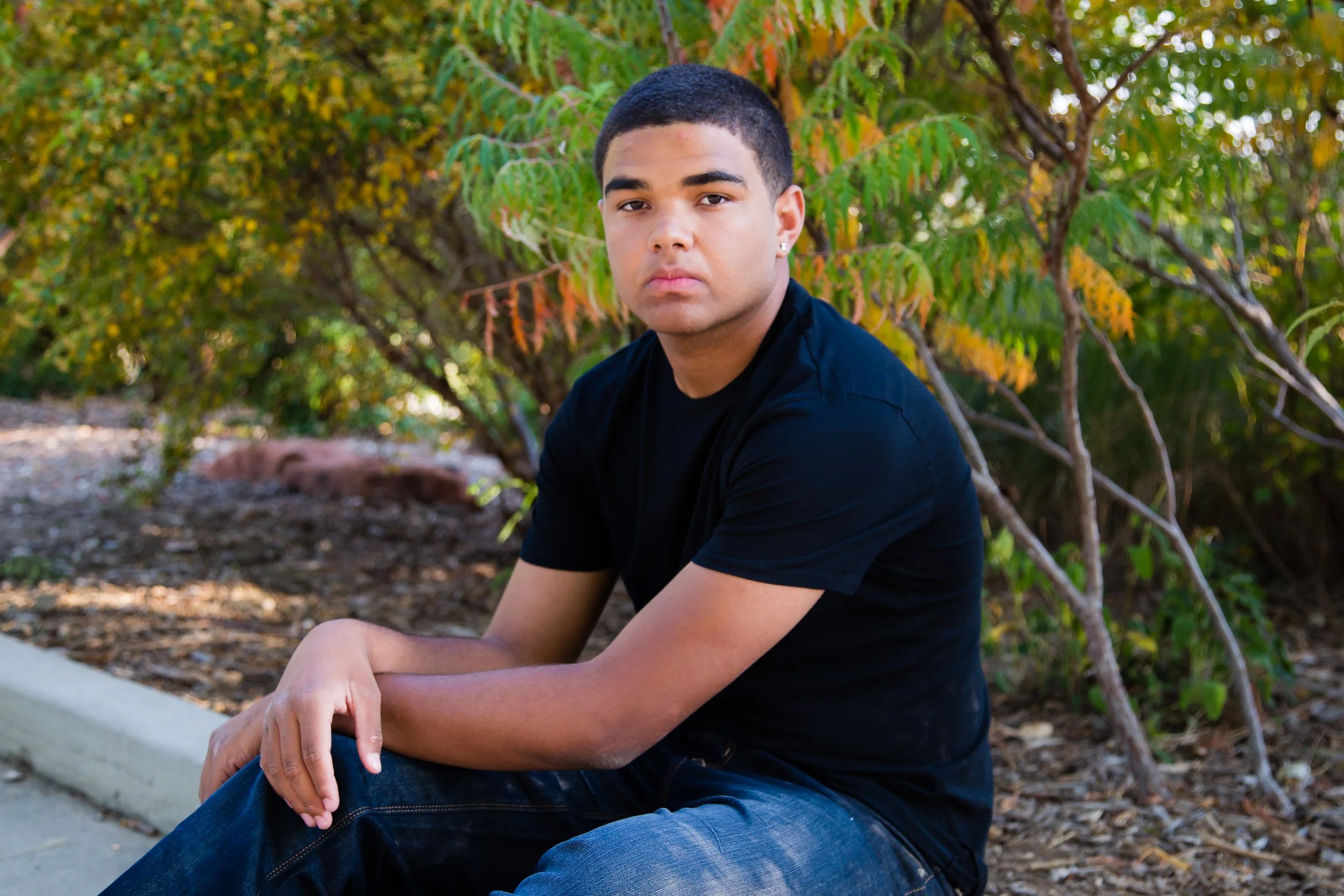 Young man with short black hair and earrings sitting outdoors near trees with green and yellow leaves.