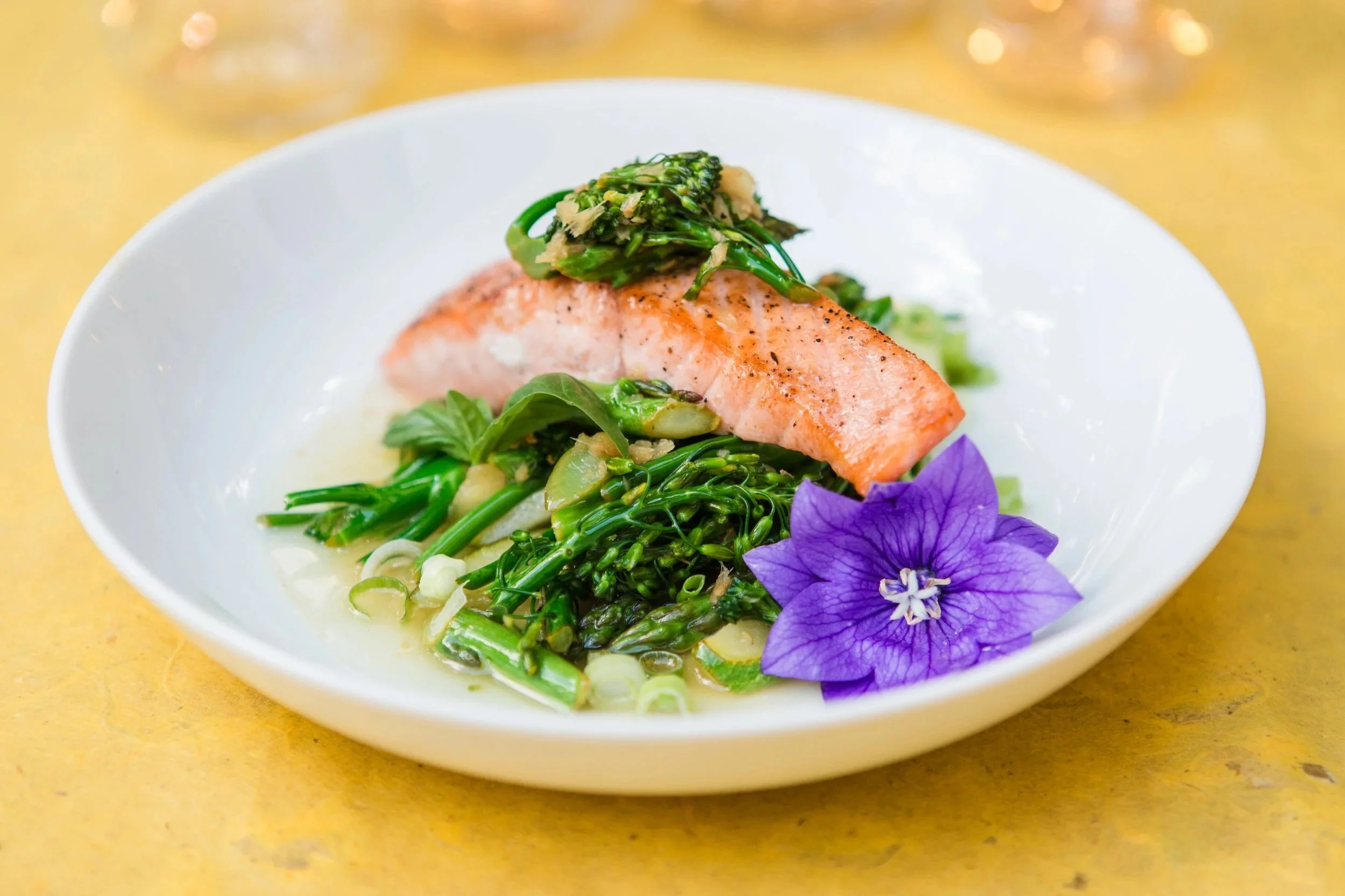 A white bowl containing cooked greens, a piece of cooked salmon, and garnished with a purple flower.
