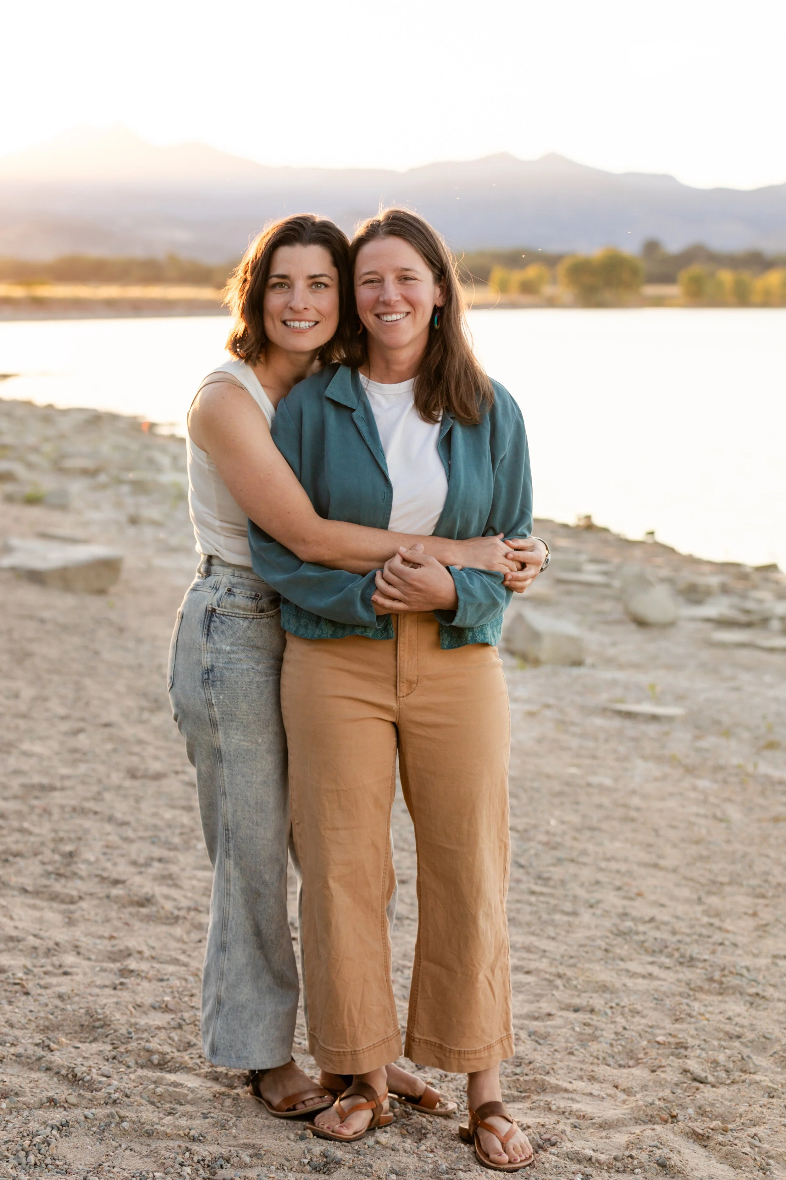Two women standing on a rocky beach during sunset, with mountains in the background. They are smiling, embracing, and wearing casual clothing and sandals.