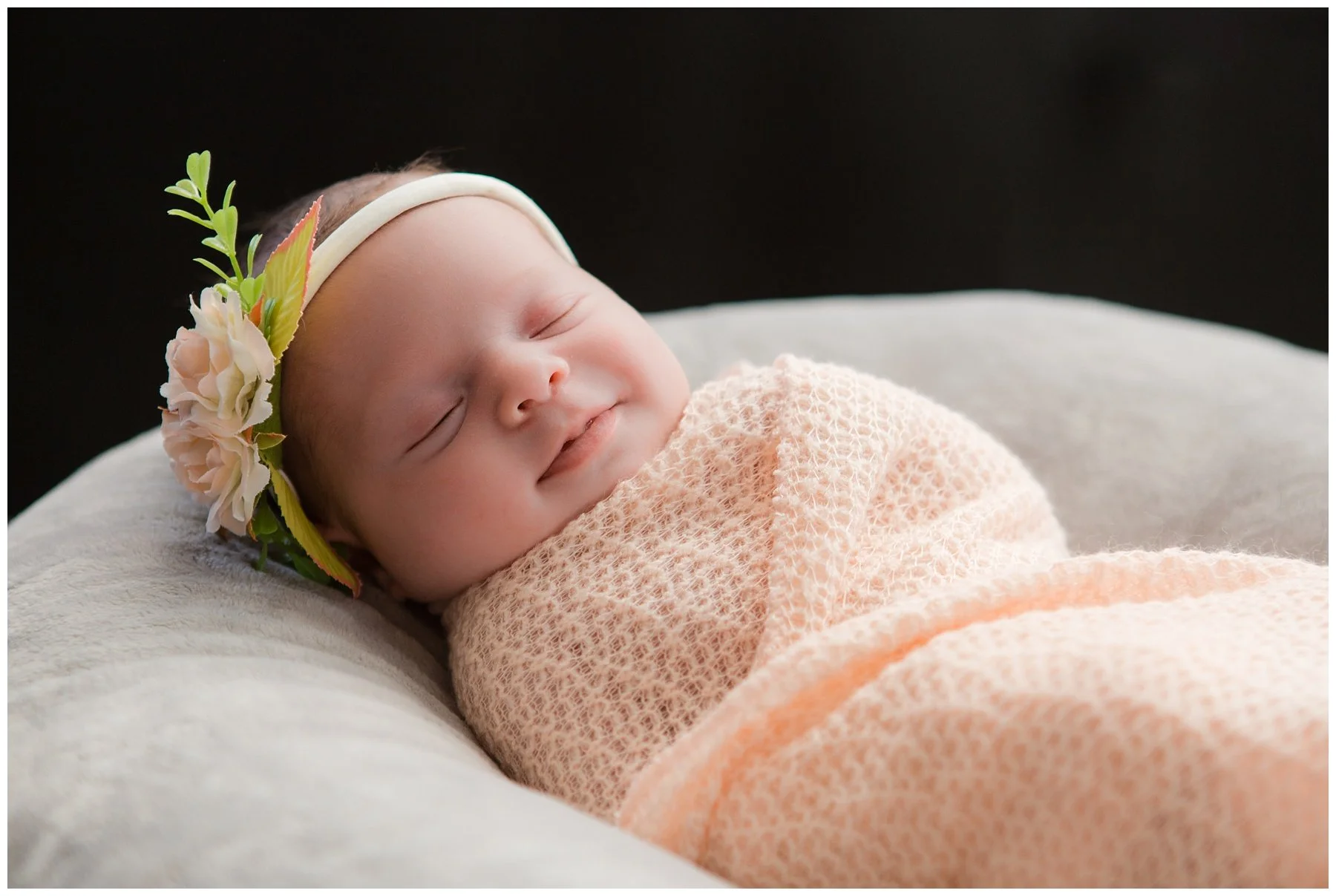 A sleeping baby girl with a floral headband, wrapped in a peach-colored blanket, lying on a soft white surface against a dark background.