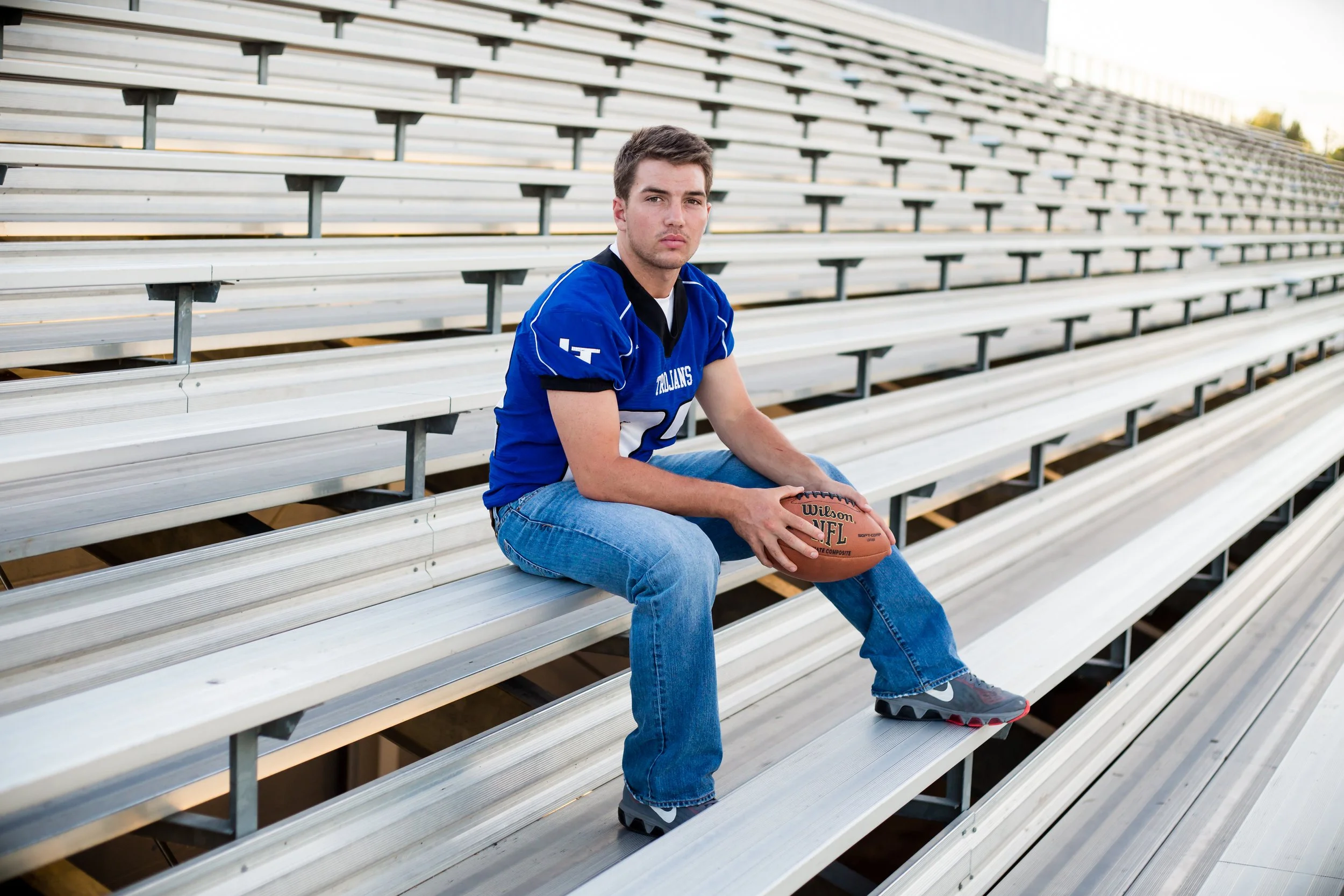A young man sitting on stadium bleachers holding a football, wearing a blue sports jersey with white accents, jeans, and athletic shoes, outdoors at the stadium during daylight.