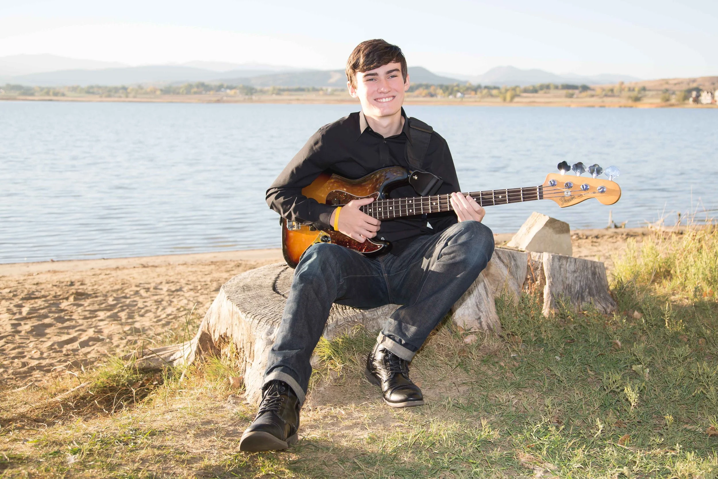 A personalized high school senior lifestyle portrait session featuring a young man sitting on a tree stump by the water with his electric guitar at McIntosh Lake in Longmont, Colorado, by Lisa Doane Photography.