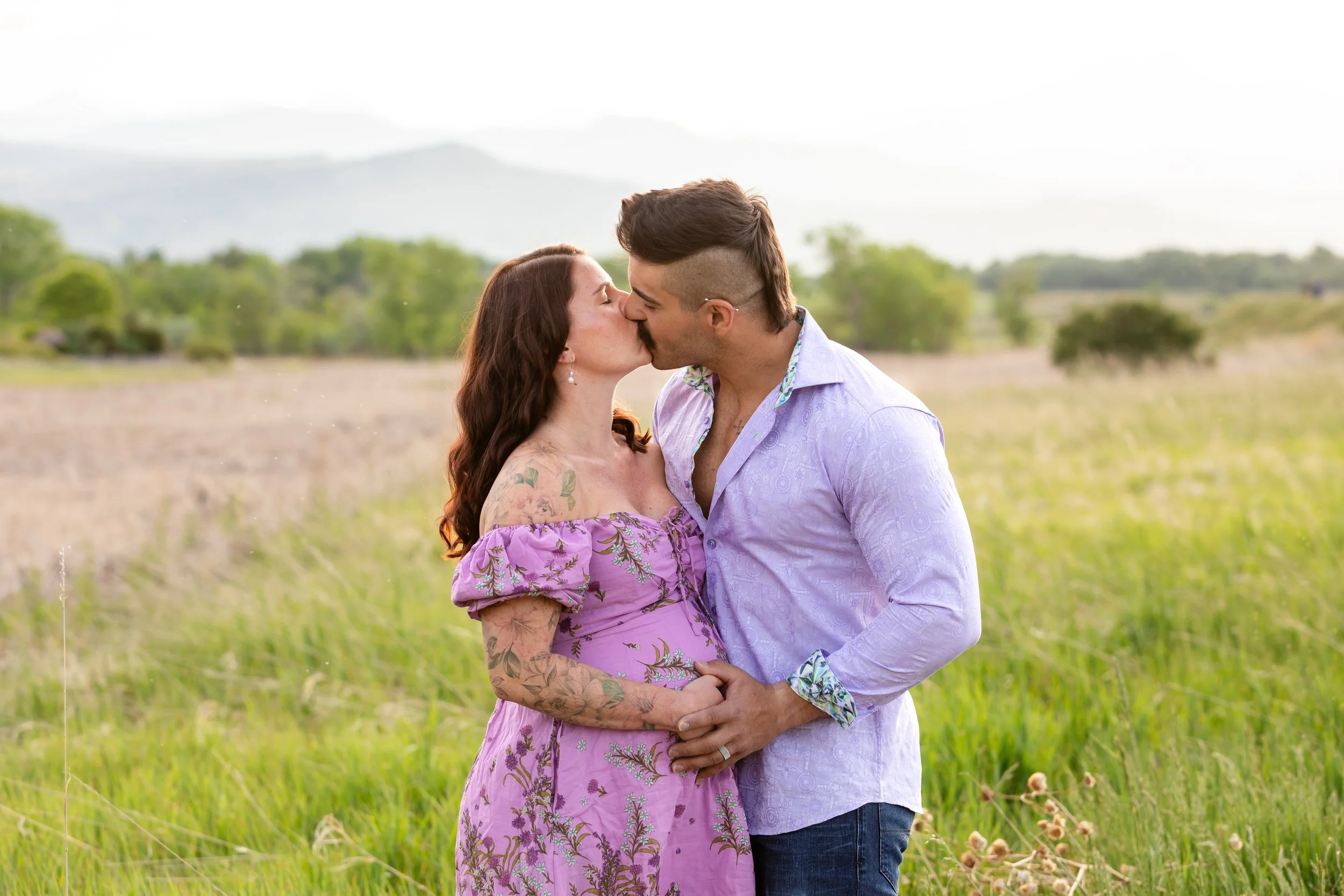 Outdoor maternity portrait session at McIntosh Lake in Longmont featuring a couple kissing in a green field with the foothills blurred in the background.