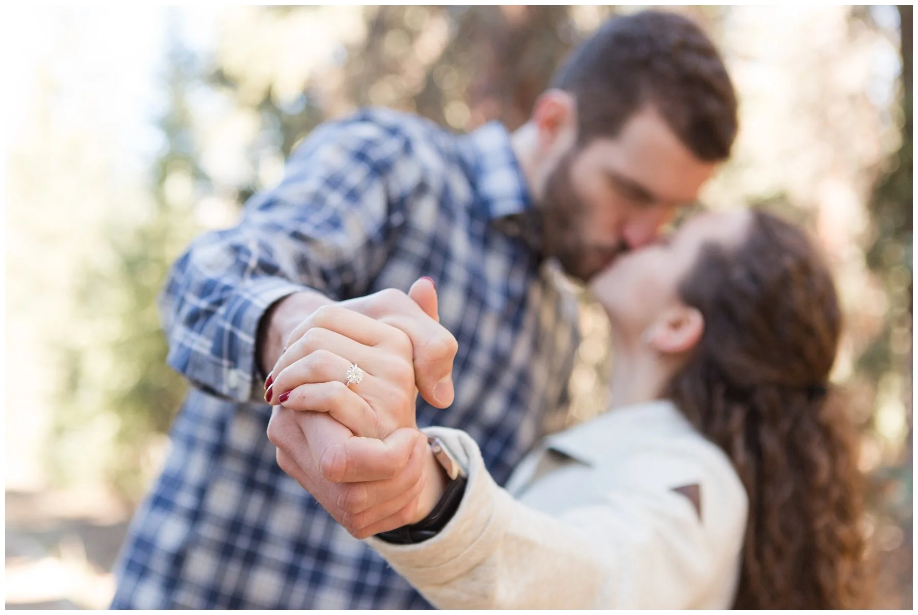 A couple dancing outdoors, holding hands with a focus on the woman's engagement ring, with their faces close together in a blurred background of trees and sunlight.