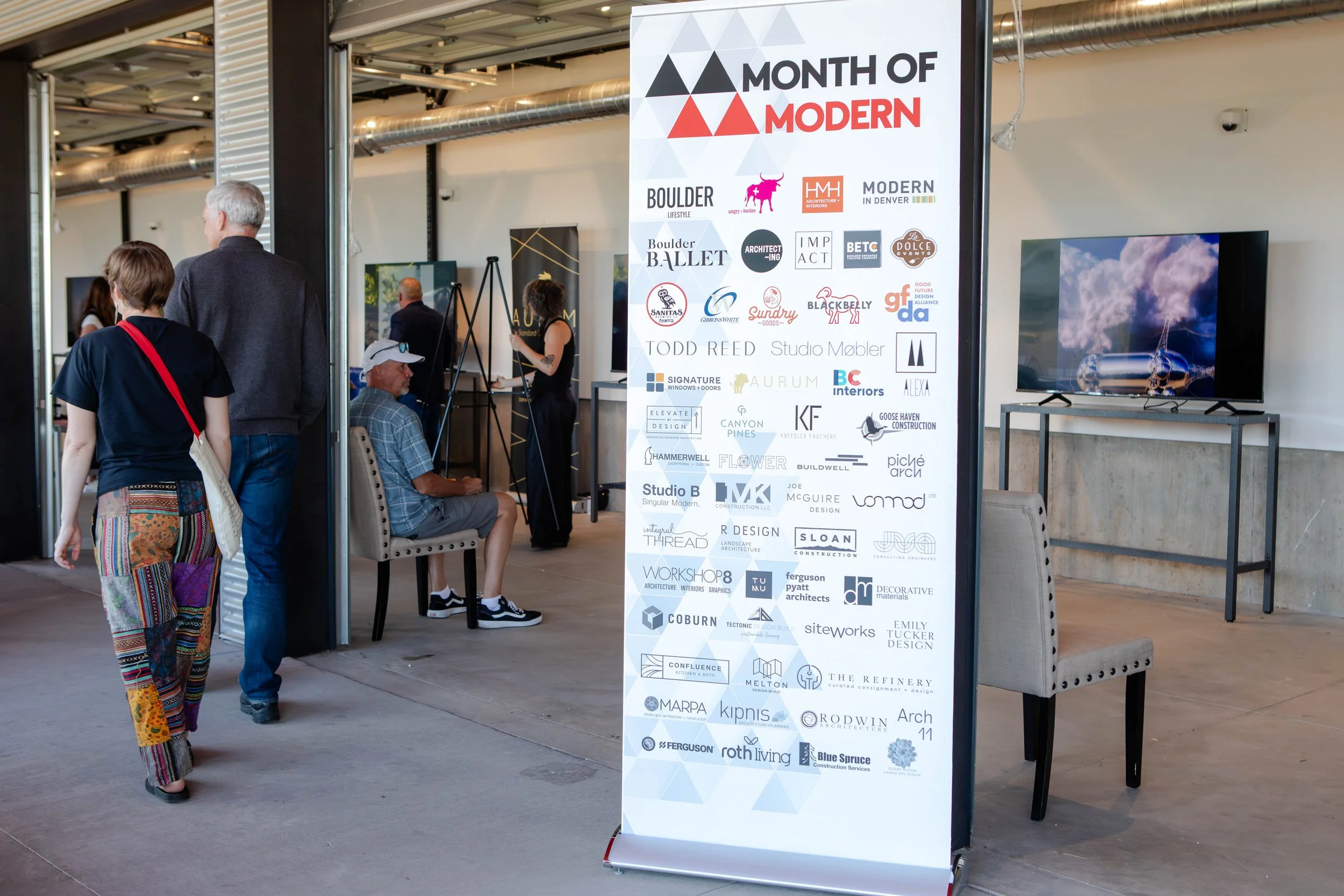 People attending an exhibition or event, looking at a large display board with numerous company logos and a screen showing an outdoor scene. The display banner reads 'Month of Modern' and features various business and design related logos.