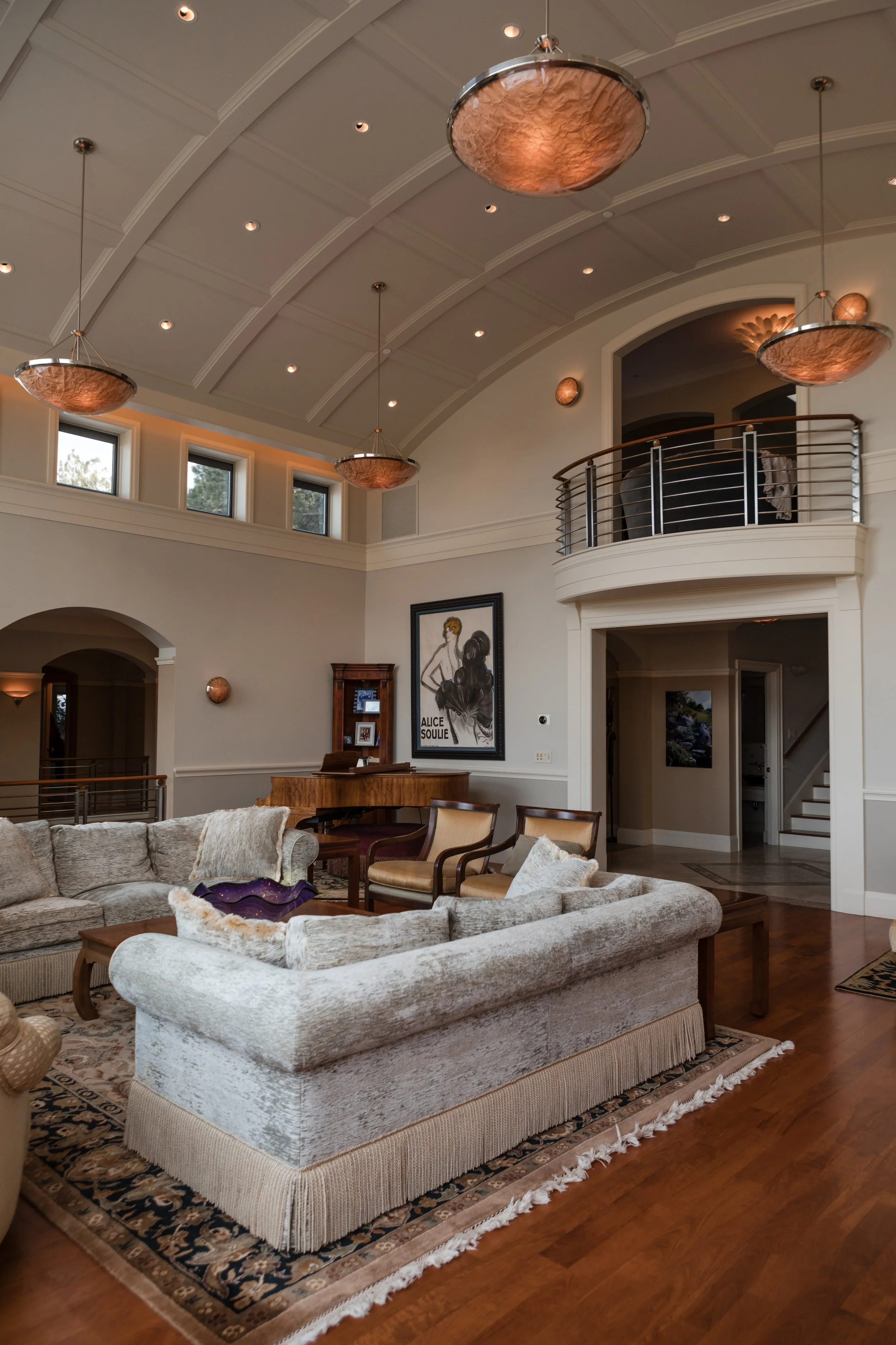 Interior of a spacious living room with high vaulted ceiling, pendant lighting fixtures, a leather sofa, armchairs, a rug, and artwork on the walls. There is also a balcony with a metal railing.