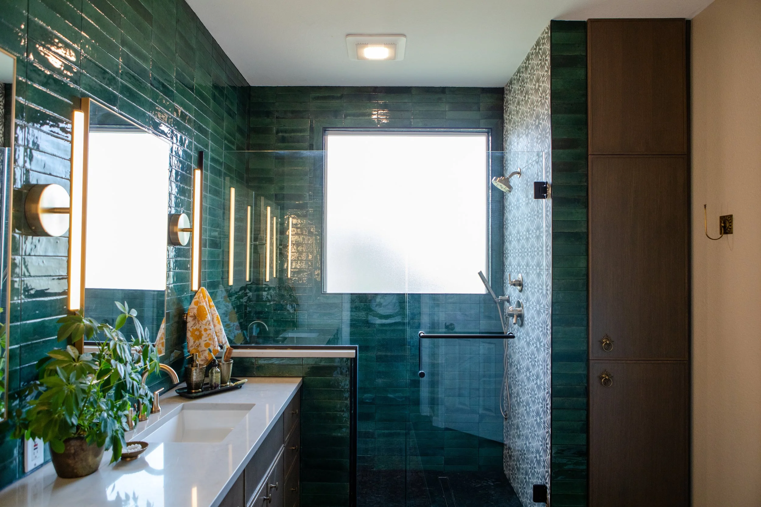 Bathroom with green tile walls, frosted window, shower area with glass divider, wooden cabinets, and a white countertop with potted plants and toiletries.