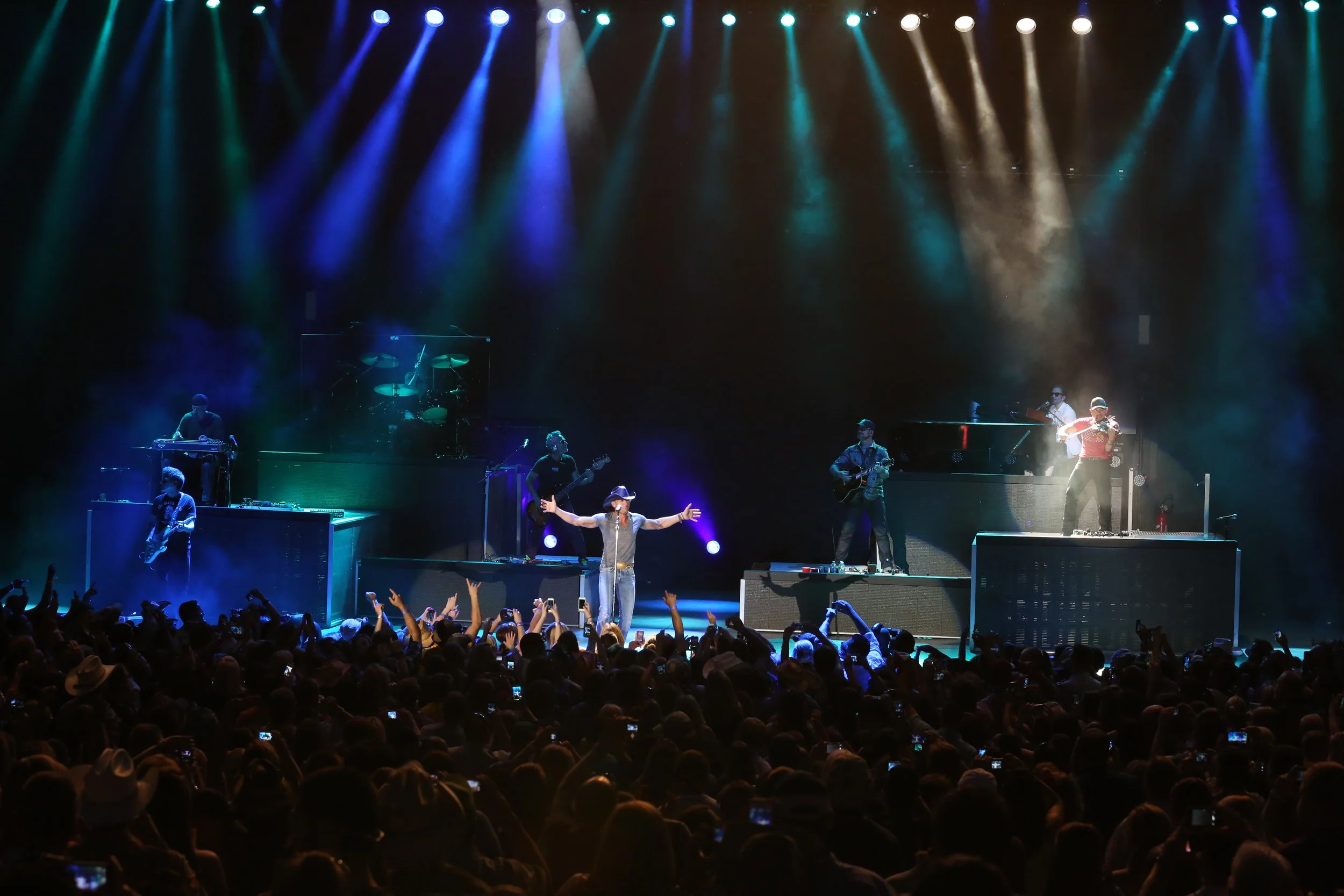 A live concert on a large stage with multiple musicians performing. There are colored lights shining down, and the audience is visible in the foreground with many raising their hands, cheering, and some taking photos or videos.